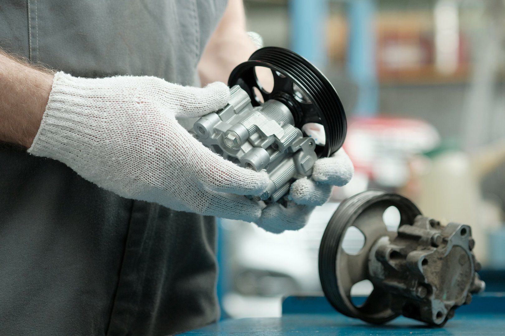 An auto mechanic is preparing to replace the hydraulic power steering pump. A new rotary oil pump is in the hands of an auto mechanic. There is a faulty pump on the worktable.