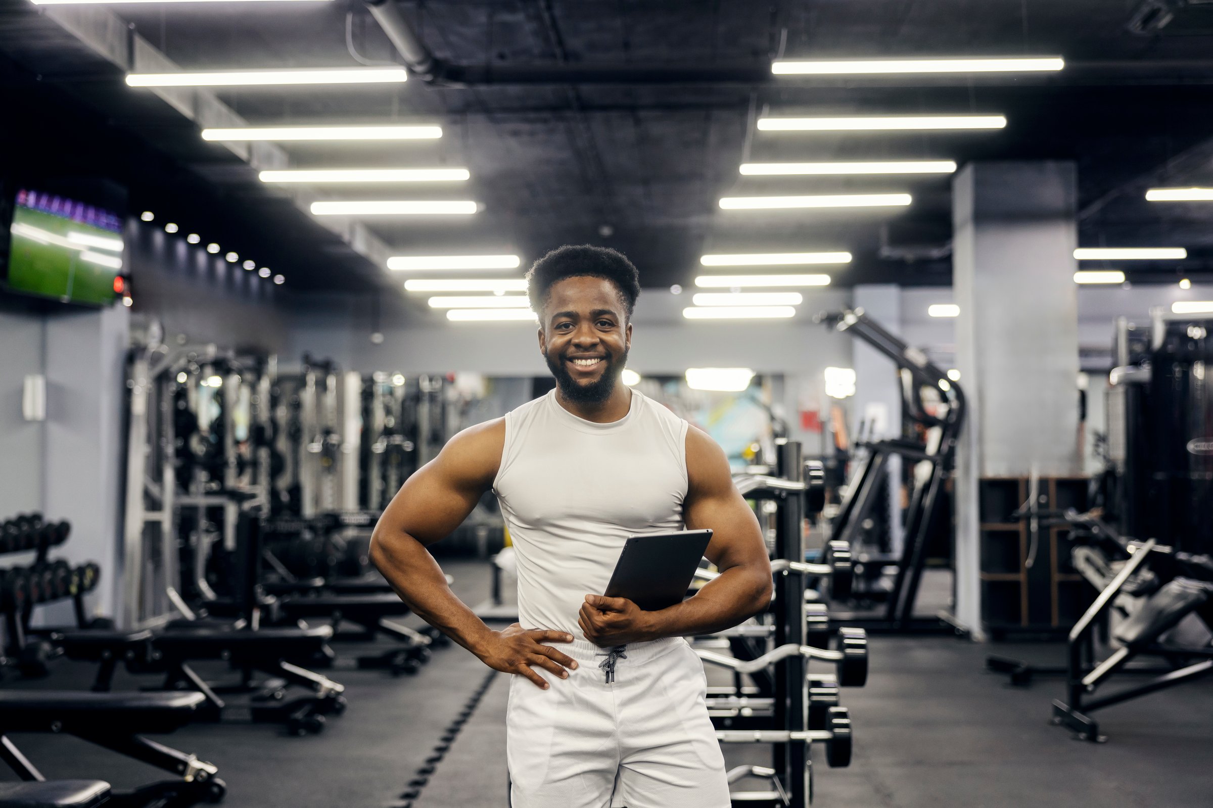 Male personal trainer holding a digital tablet and smiling confidently in a modern gym