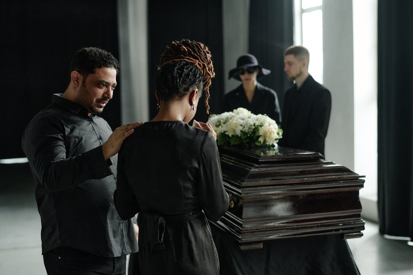Black woman standing at coffin being comforted by Caucasian man during funeral ceremony, two young adults in black clothing standing in background near window, floral arrangement on casket