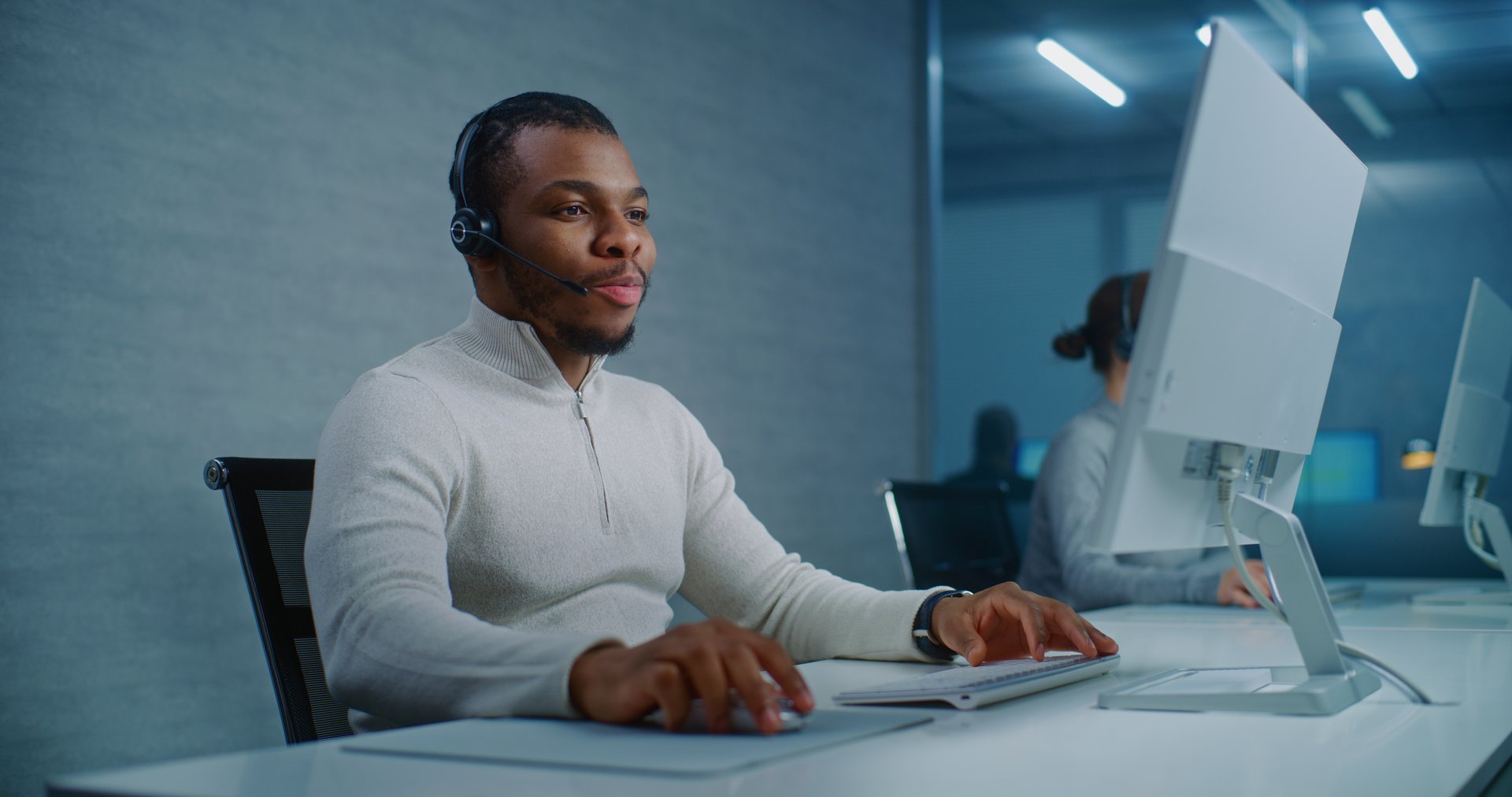 Call center office: African American hotline operator talks on headset with customer, works on computer, provides online assistance for client. Diverse team of technical support specialists working.