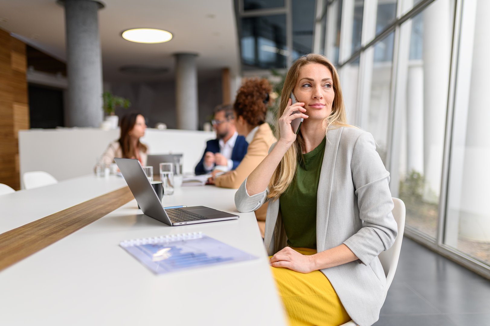 Blond entrepreneur discussing over phone call while sitting in board room with professional teammates in the background