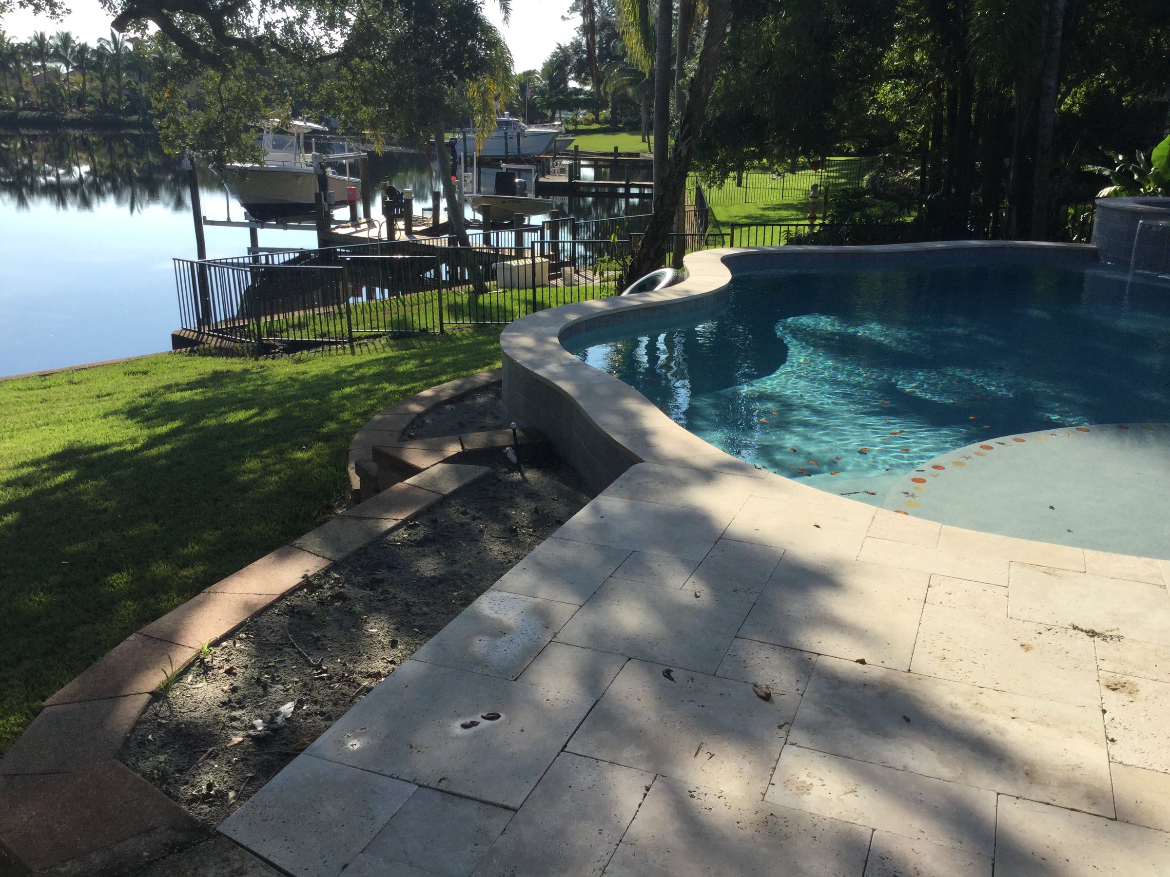 Backyard pool with stone patio overlooking a lake and docked boats amid lush green trees on a sunny day.
