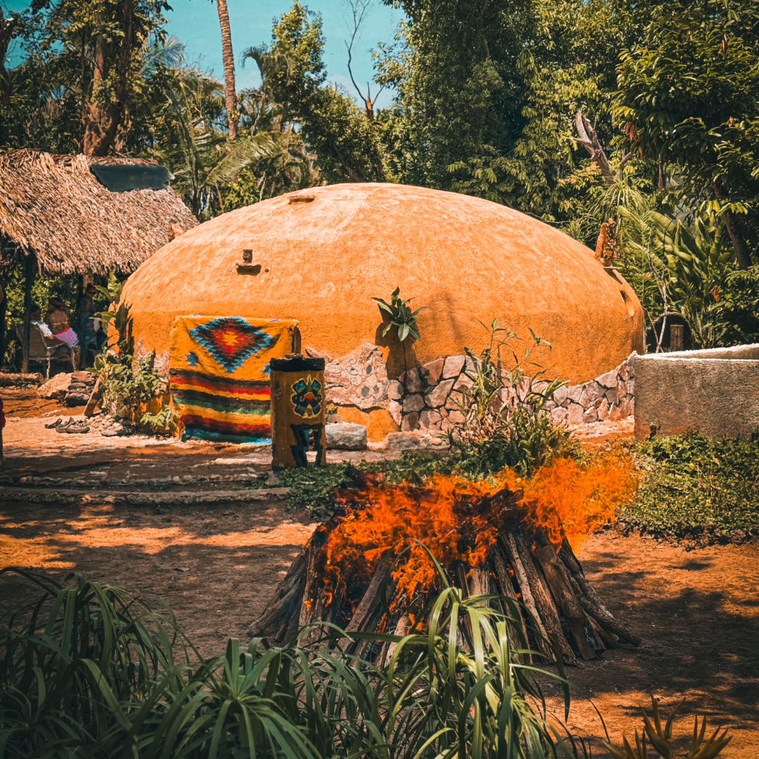Dome-shaped hut with colorful blanket, surrounded by lush greenery, and a small bonfire in the foreground.