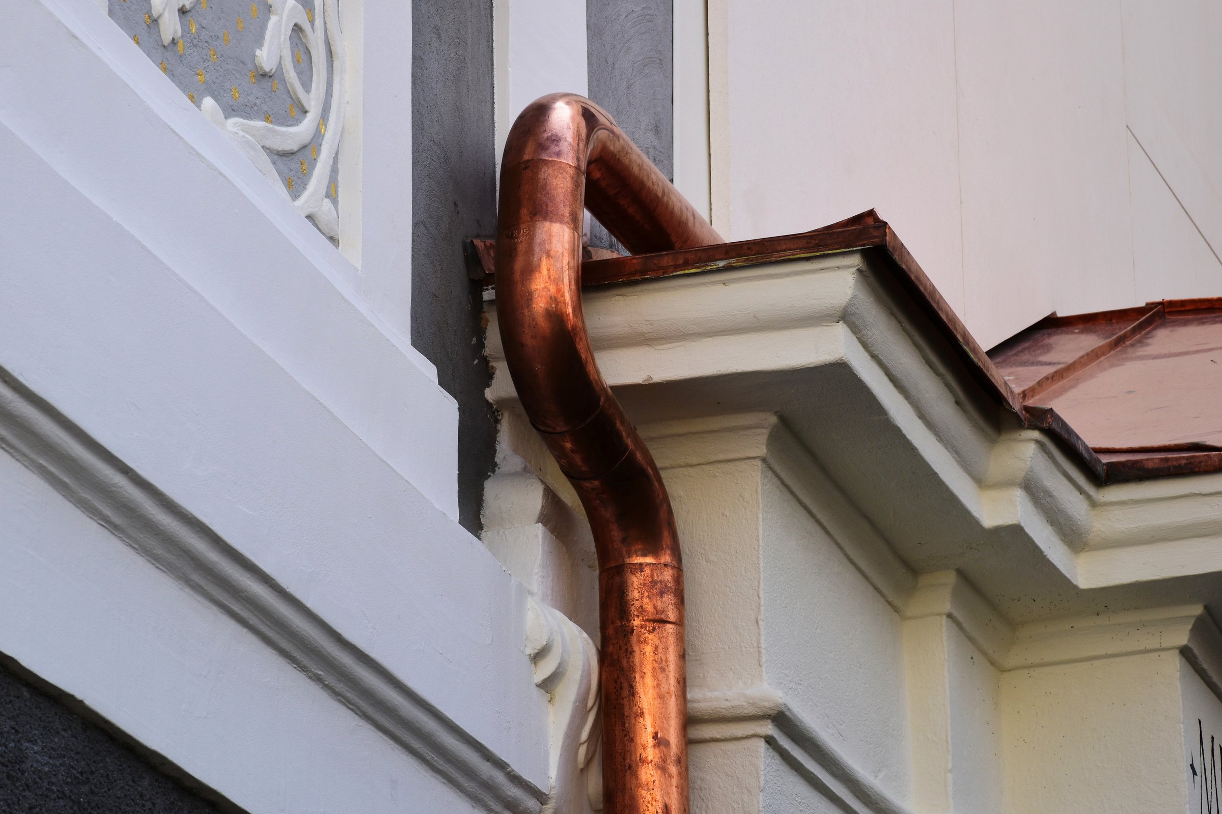 shiny new copper rain water leader, downspout or down pipe detail with elbow. white stucco elevation. home renovation, construction concept. textured white stucco exterior wall and window background. blue sky.