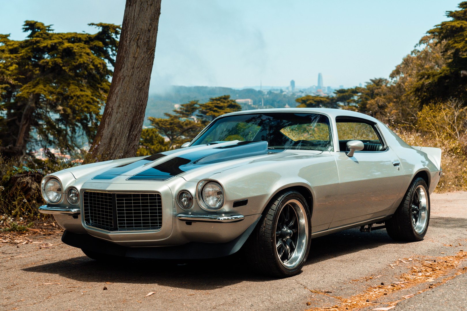 A beautifully restored vintage Camaro stands proudly against the scenic backdrop of San Francisco, blending classic American muscle with the city's iconic charm. The shimmering silver finish and bold racing stripes evoke a sense of nostalgia and power, making this a perfect fusion of style and strength.