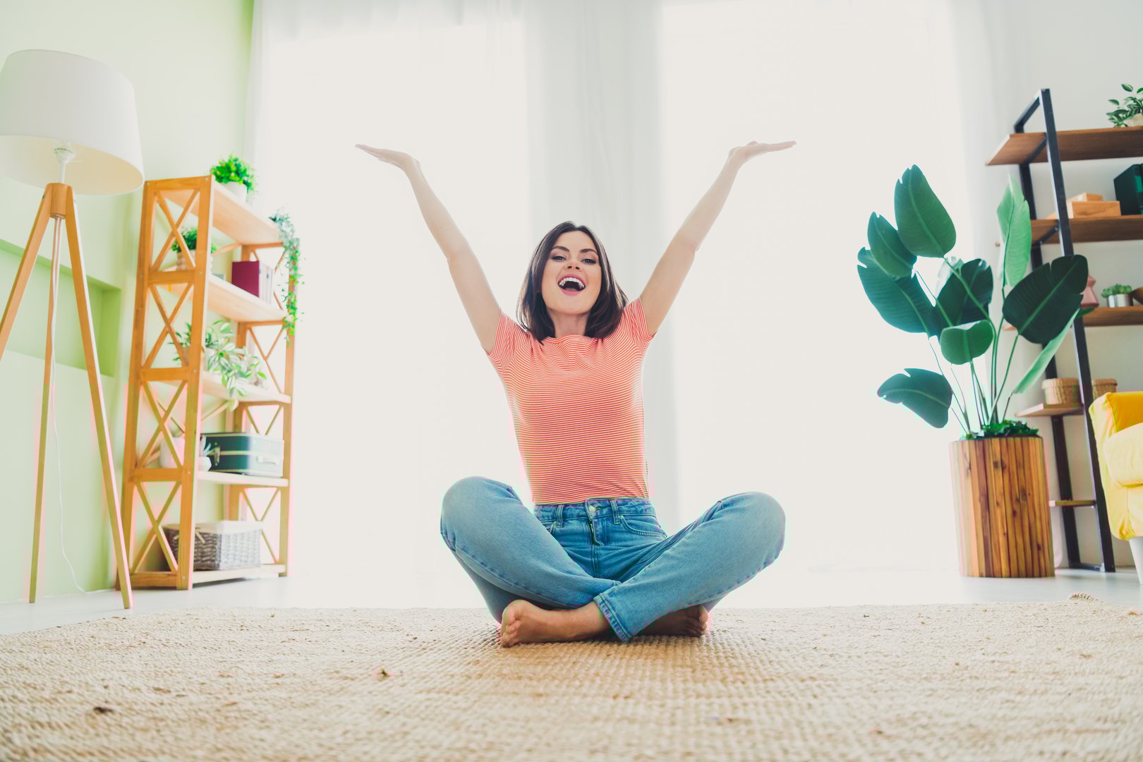 A cheerful young woman sits cross-legged in a bright, cozy living room, enjoying a casual weekend indoors. The space is stylish and filled with natural light.