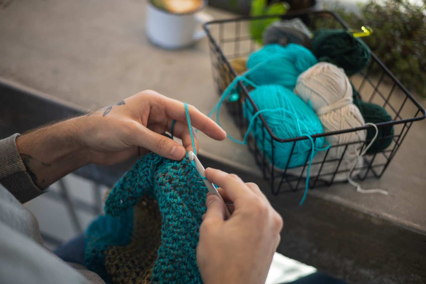 Male hands crochet handmade artwork use wool yarn and hook over table with coffee and potted plant top view closeup. Feminine man enjoy art hobby needlework handcrafted DIY thread sewing at home