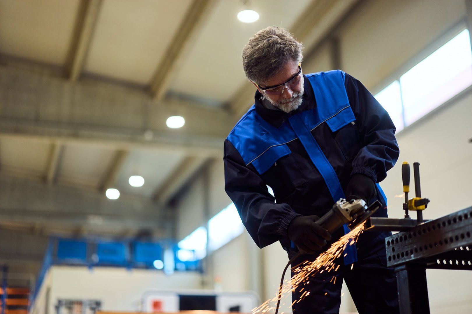 Man in protective gear working with grinder tool, creating sparks, inside factory.