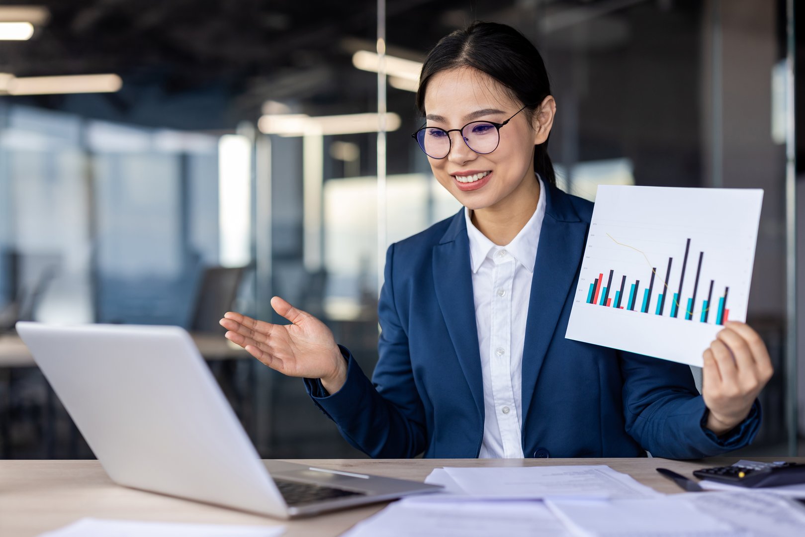 Professional woman in a business suit shows a colorful bar graph chart while video conferencing on a laptop.