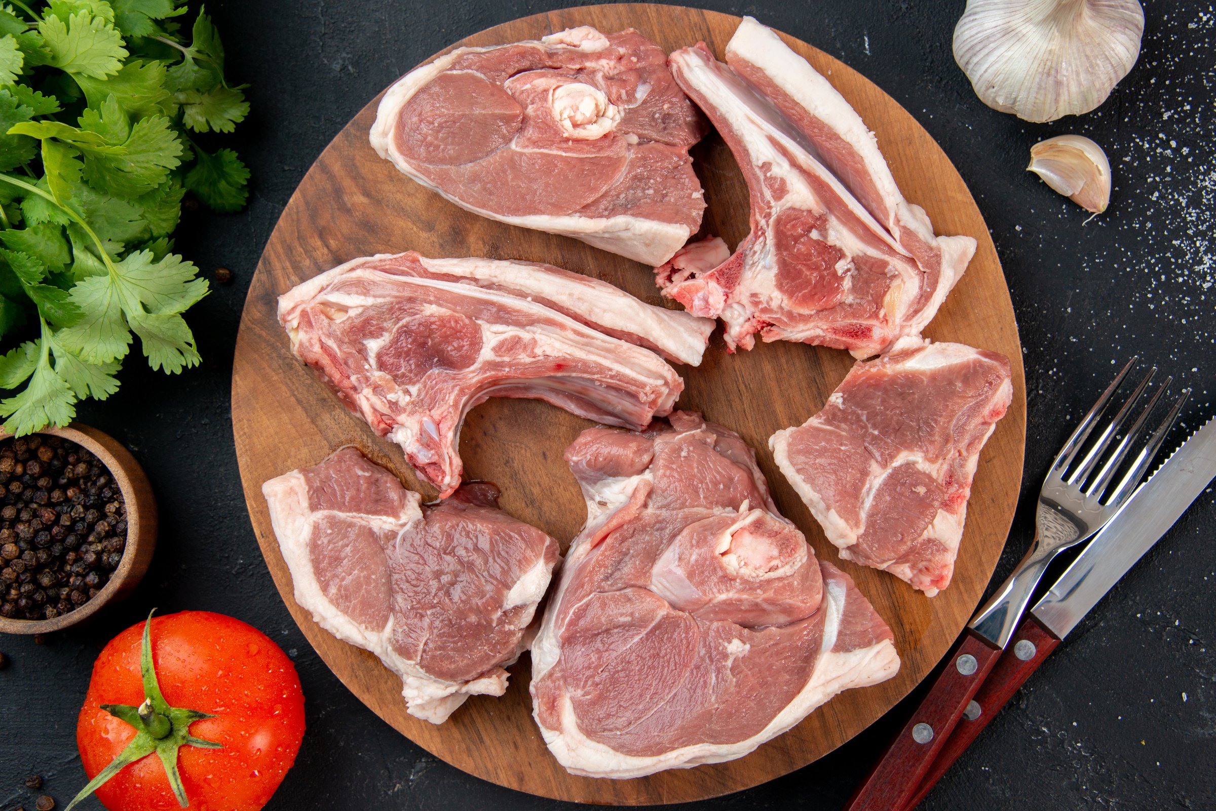 Raw lamb chops on a wooden board surrounded by garlic, tomato, cilantro, peppercorns, salt, and cutlery on a dark background.