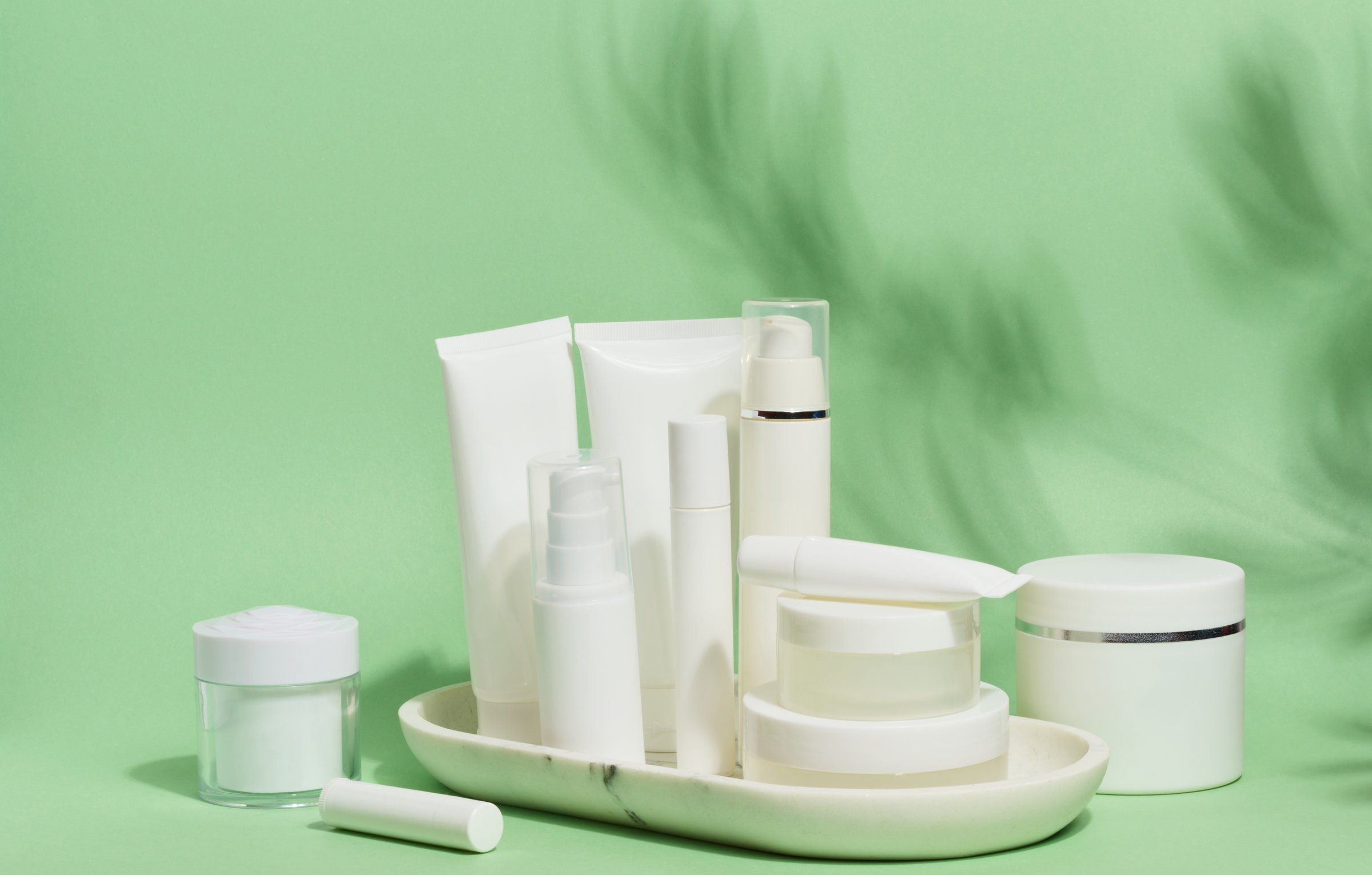 A collection of various white cosmetic containers, including tubes, jars, and bottles, are arranged on a white stand against a light green background with leaf shadows.
