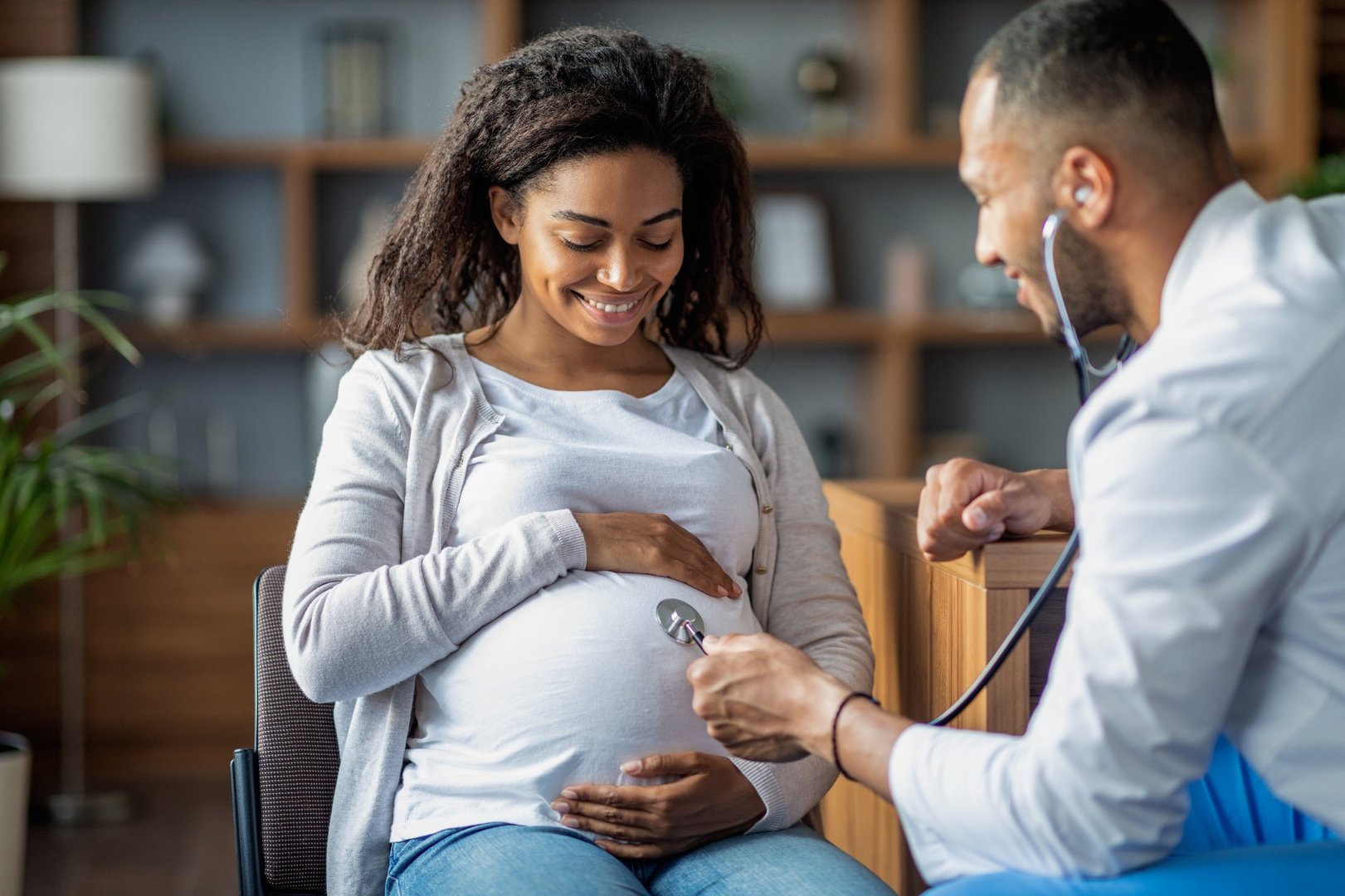 Pregnant millennial black woman sitting on chair at clinic, touching her big tummy and smiling, visiting doctor, arab man gynecologist checking baby heart rate with stethoscope, copy space