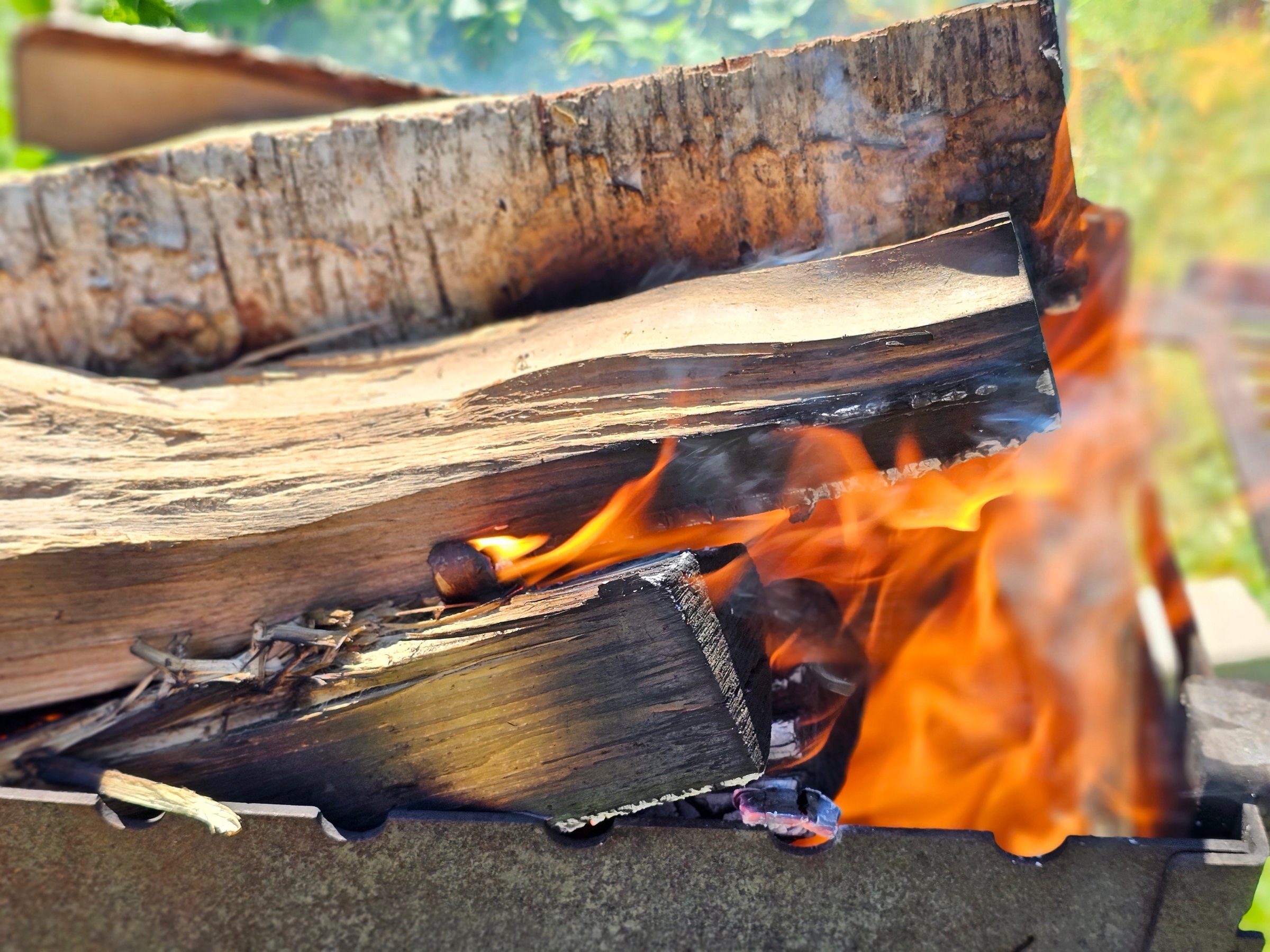 Flames dance around stacked firewood in a backyard fire pit as sunlight filters through nearby trees.