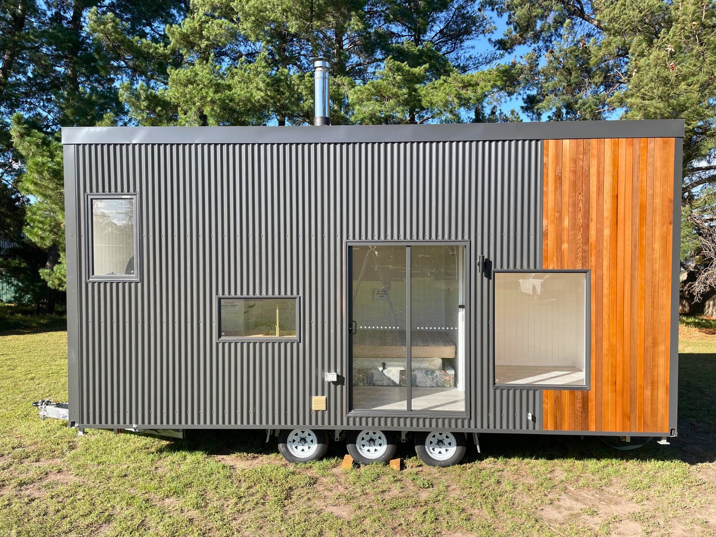 Modern tiny house with wooden and black metal facade, surrounded by greenery, lit by soft evening sunlight.