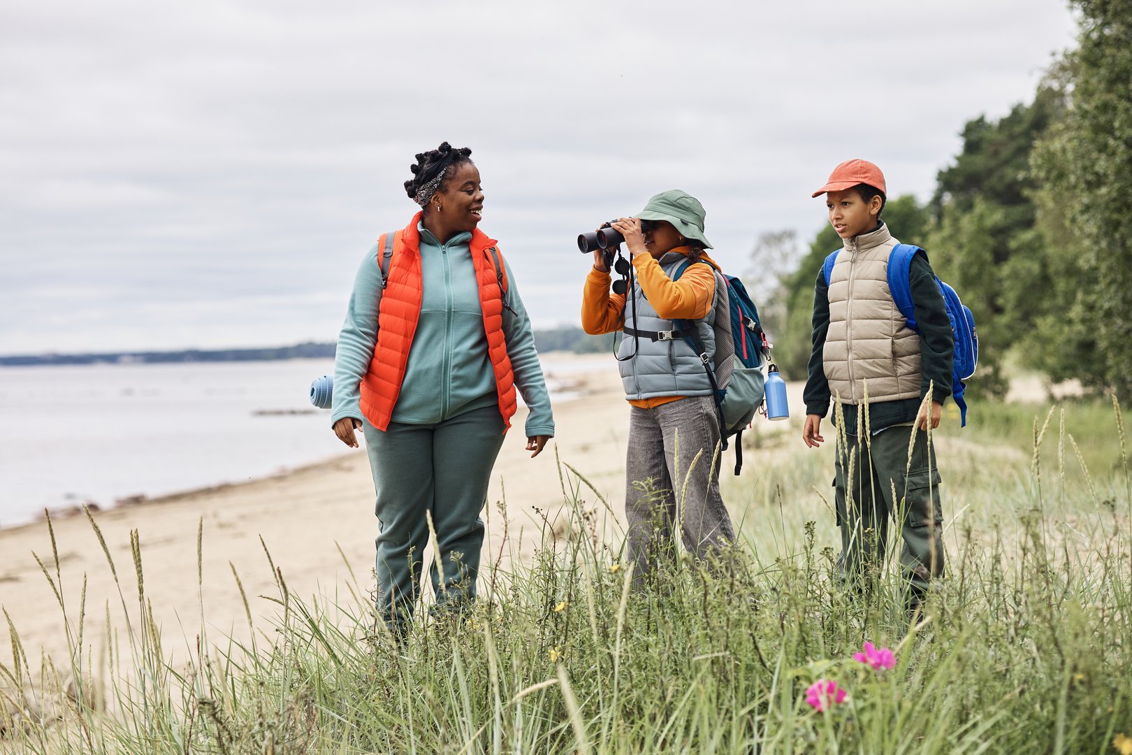 Family hiking along sandy beach, Black woman talking while boy and girl standing nearby, girl in middle using binoculars, all carrying backpacks