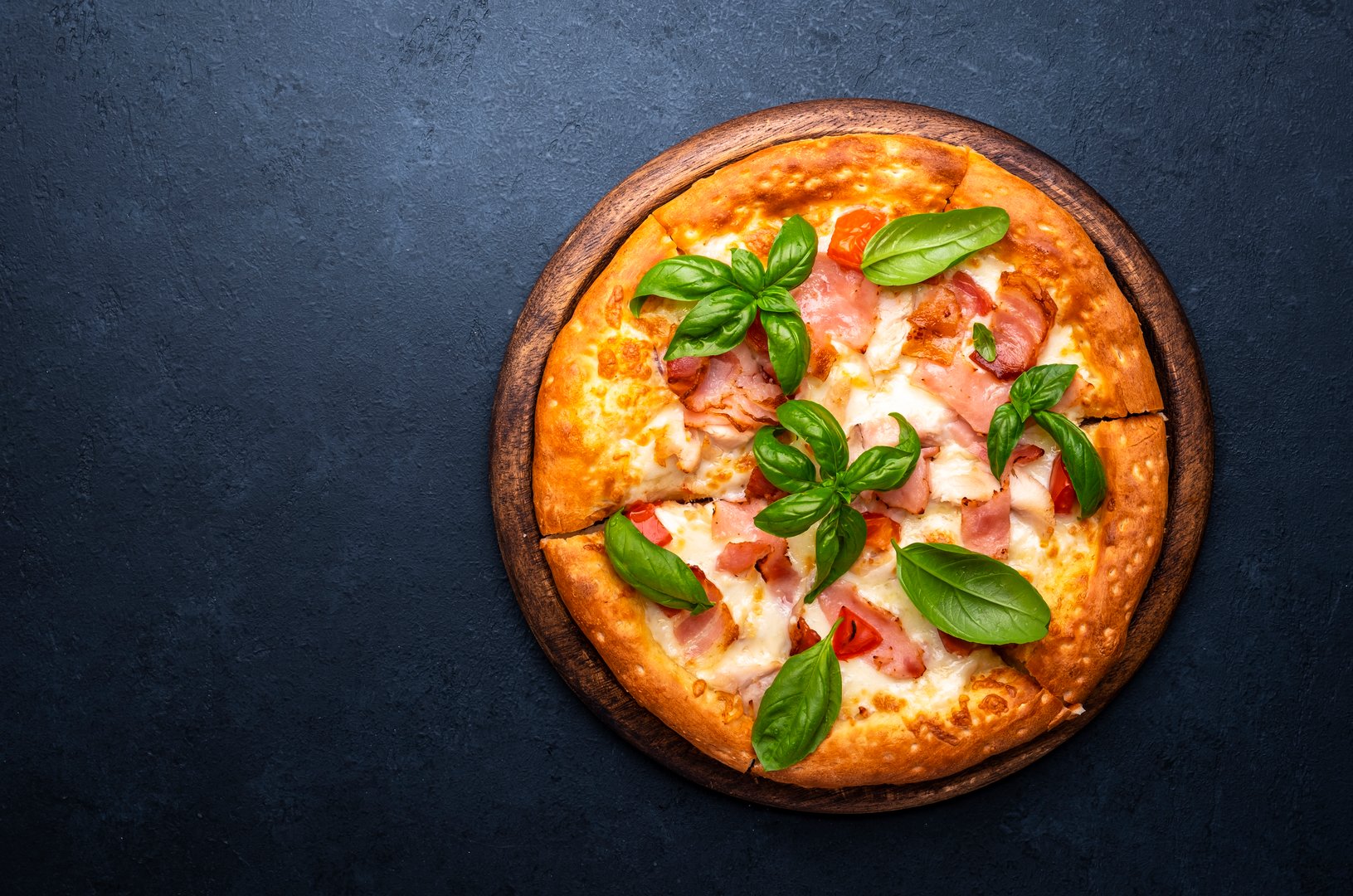 Homemade hot pizza with ham, mozzarella cheese, tomato sauce and green basil, black table background, top view