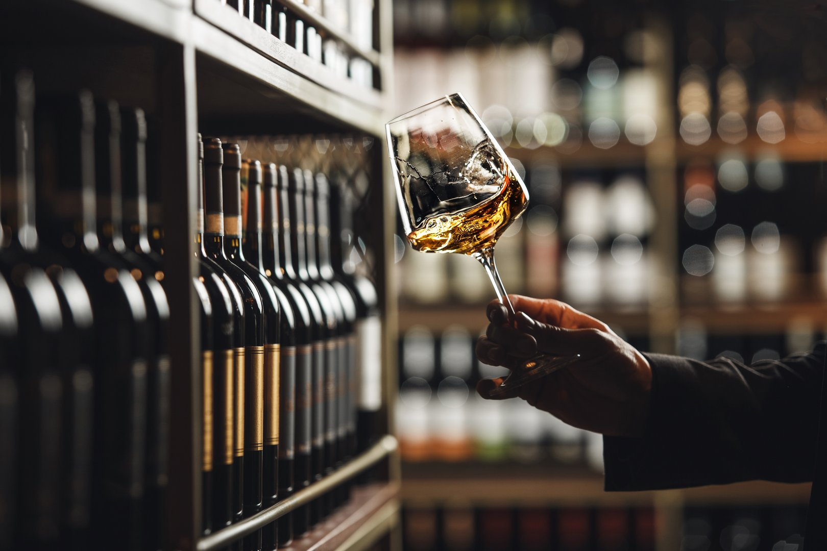 Sommelier carefully examines color and consistency of white wine held in stemmed glass, with shelves of wine bottles subtly blurred in background, suggesting an intimate, discerning setting typical for wine tasting in cellar environment.