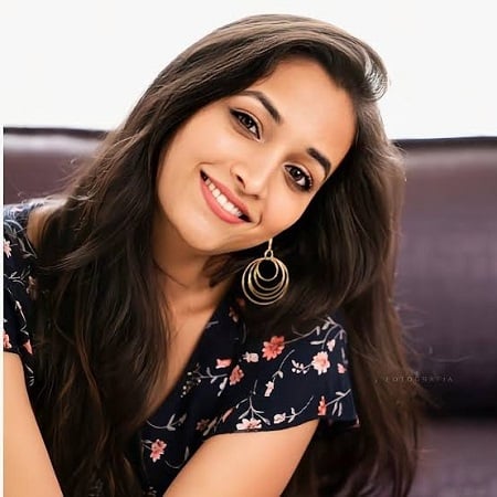 Smiling woman with long hair, wearing a floral dress and hoop earrings, sitting on a dark couch.