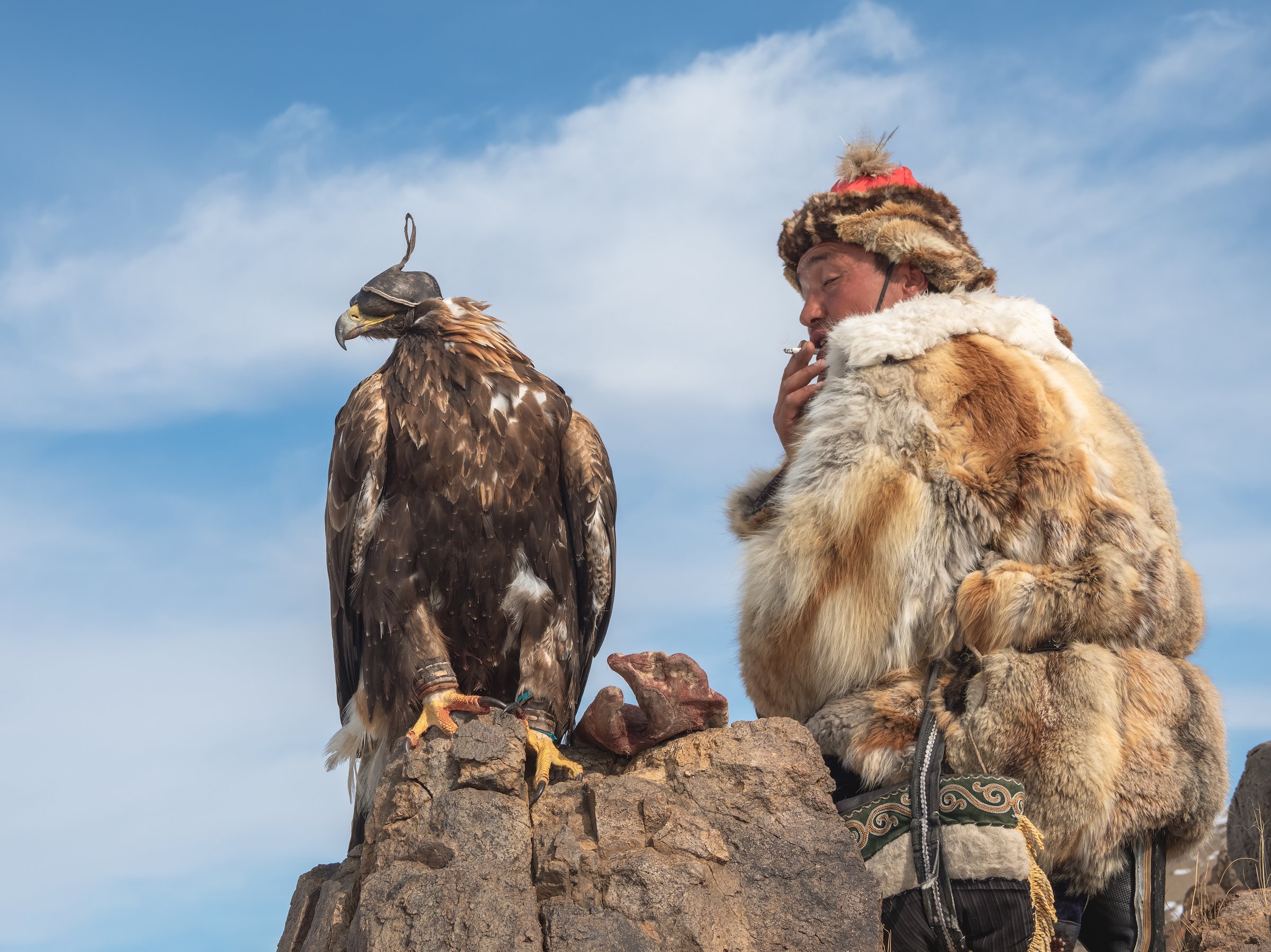 Ulgii, Mongolia - September, 30, 2023: Portrait of a smoking Mongolian hunter in winter fur clothes with his golden eagle. A sunny day in the middle of the Mongolian steppe.
