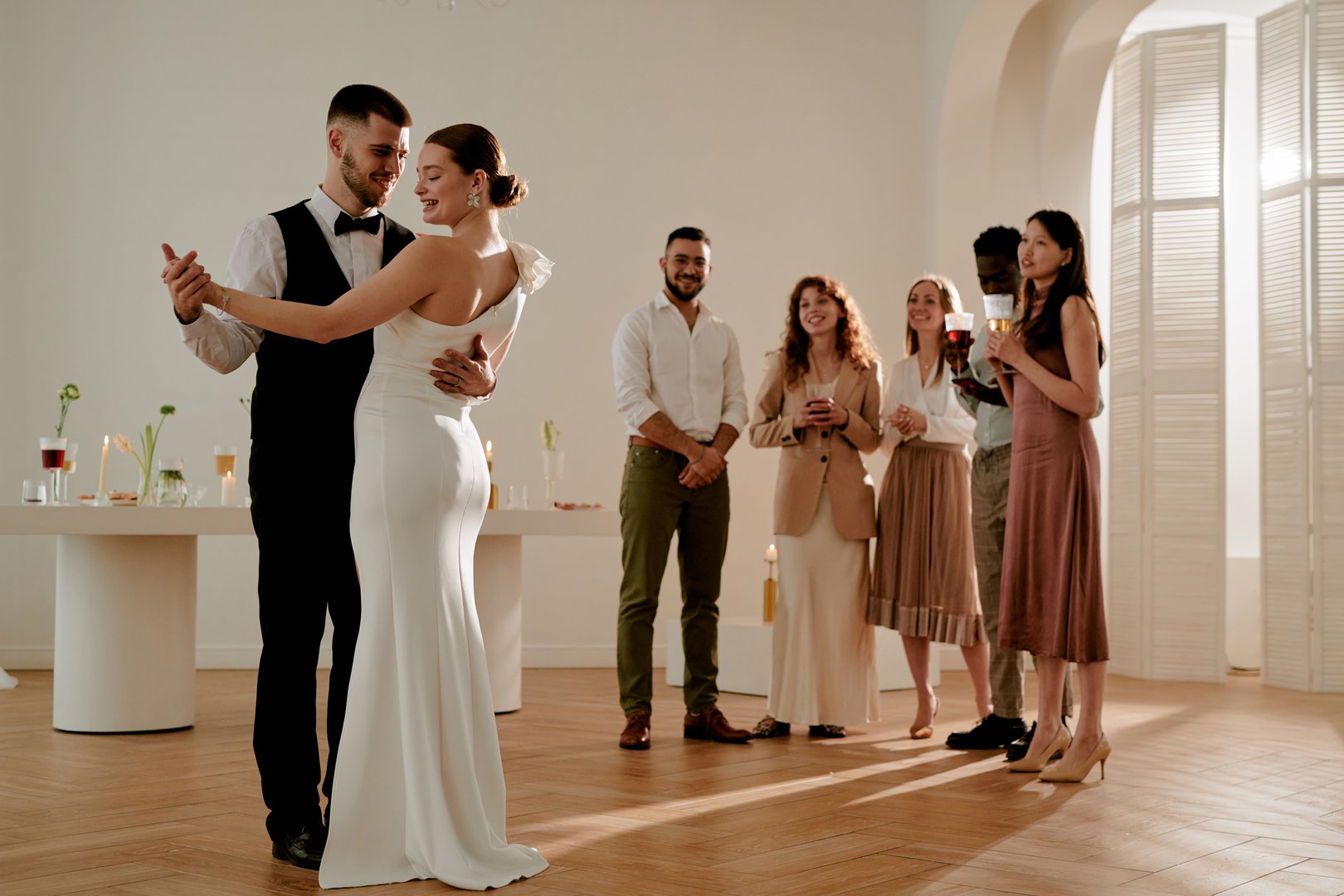 Groom and bride dancing together in wedding attire while diverse group of young adults standing in background watching and smiling