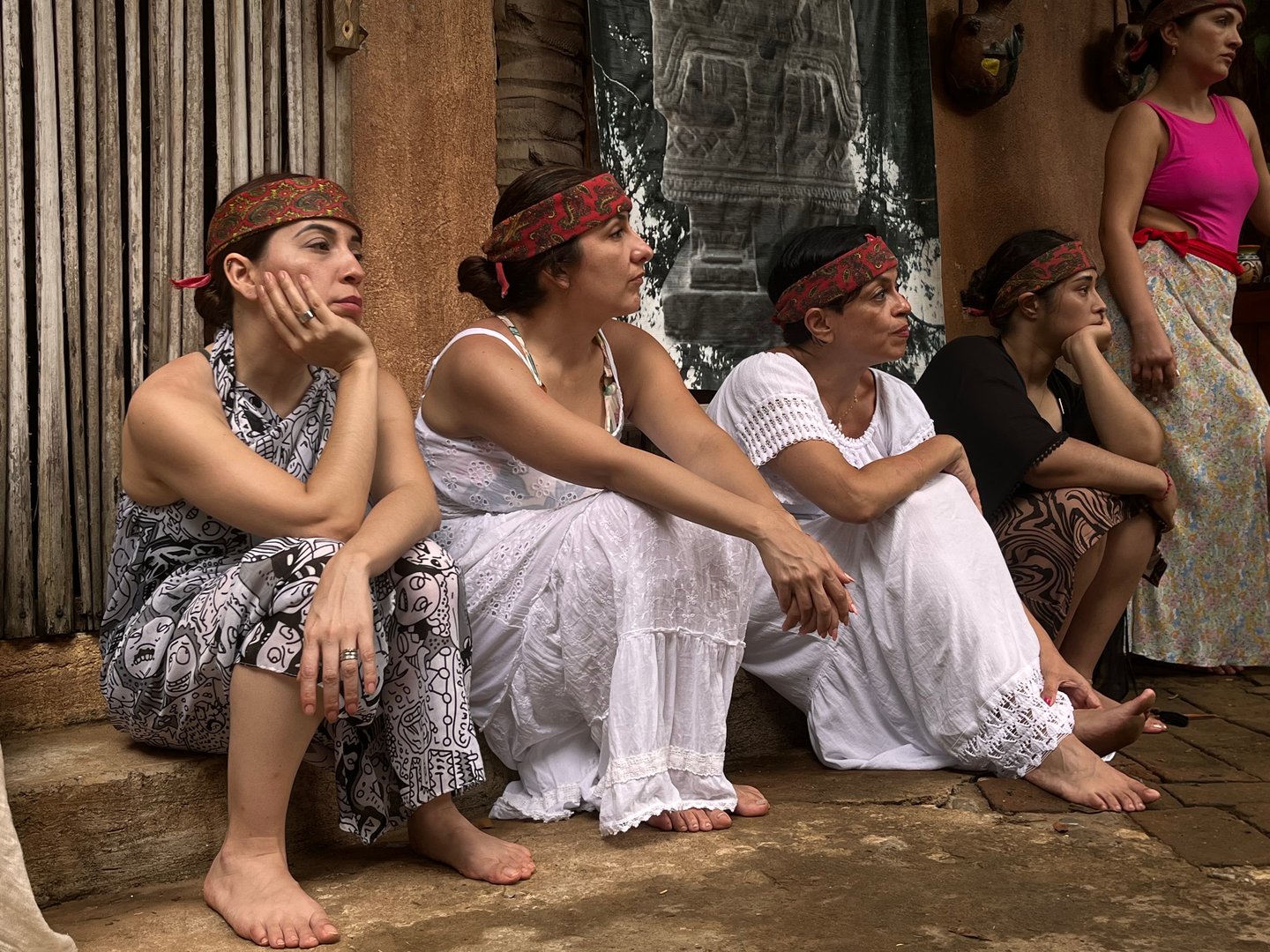Four women in patterned attire and headbands seated outdoors on a stone surface, engaged in observation or discussion.
