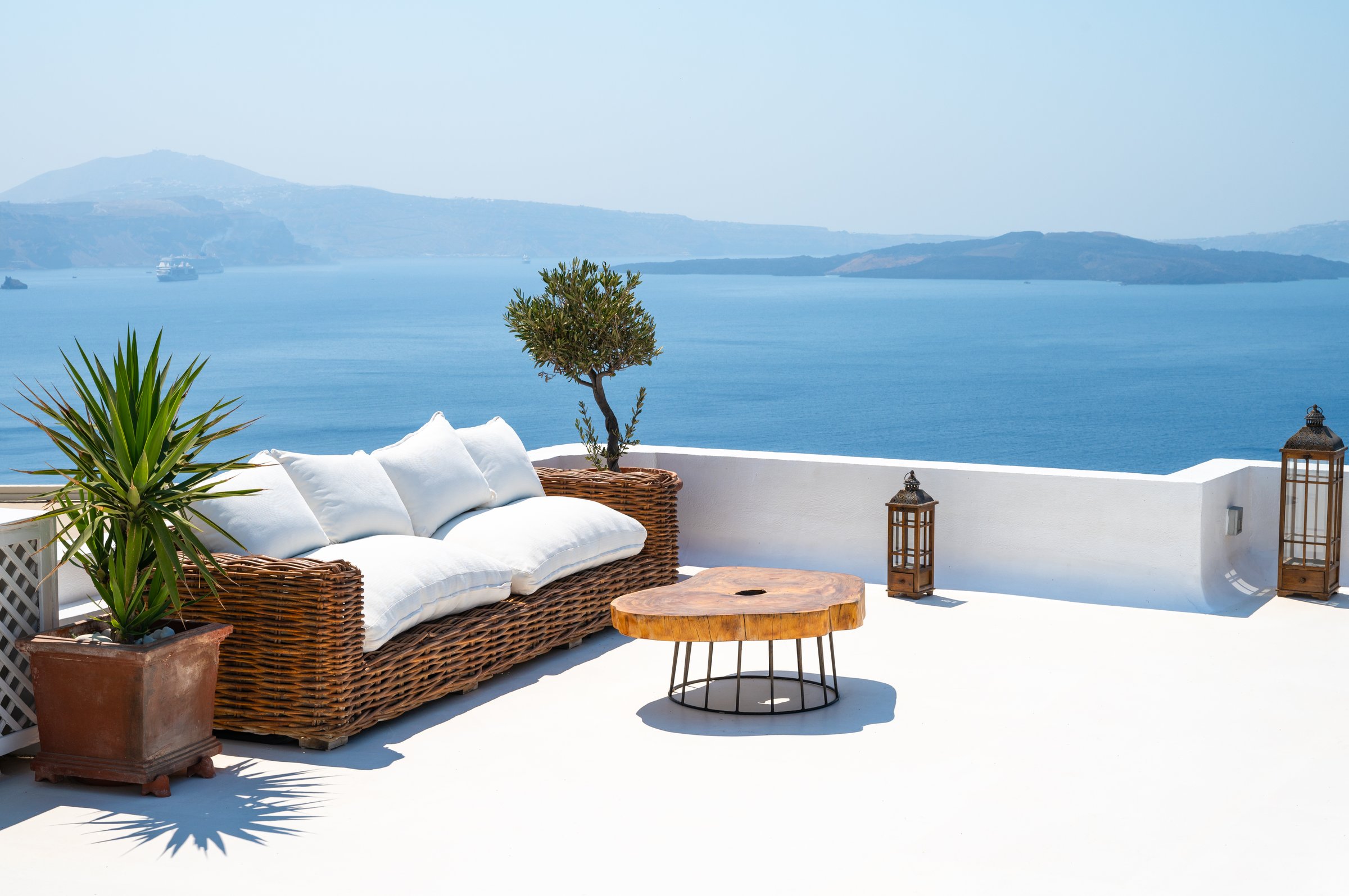Rooftop terrace with rattan seat and white pillows, stylish wooden table and potted plants on a sunny summer day in Oia village, Santorini Island, Greece. Outdoor trendy furniture.