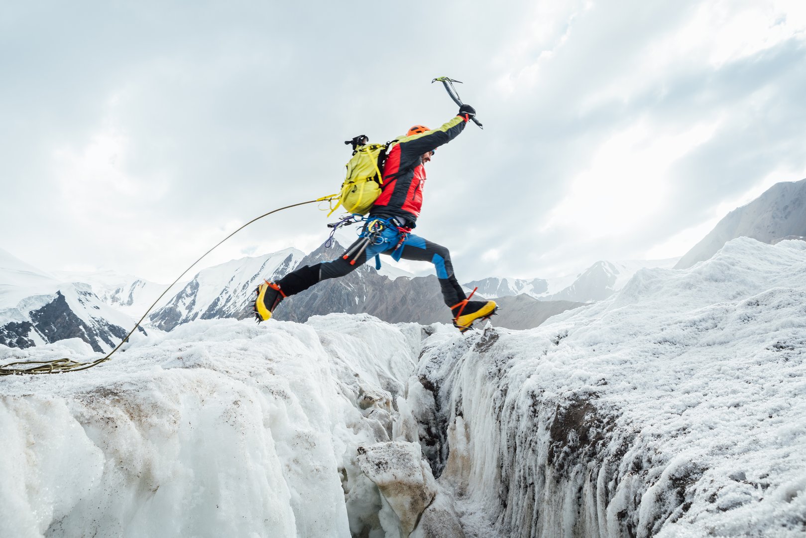 Climber in vibrant gear leaps over crevasse on icy glacier during Lenin peak ascent under clear sky with snow-covered peaks in background. Extreme active people, high-altitude mountaineering concept.