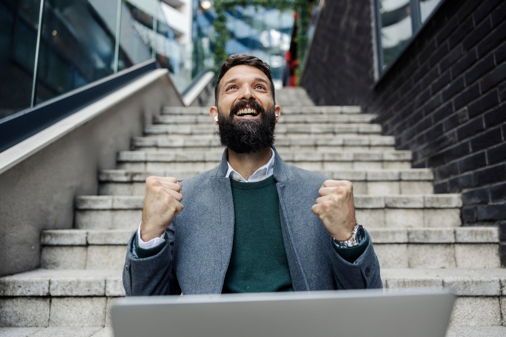 Portrait of successful elegant businessman sitting on stairs with laptop at financial district and celebrating victory.