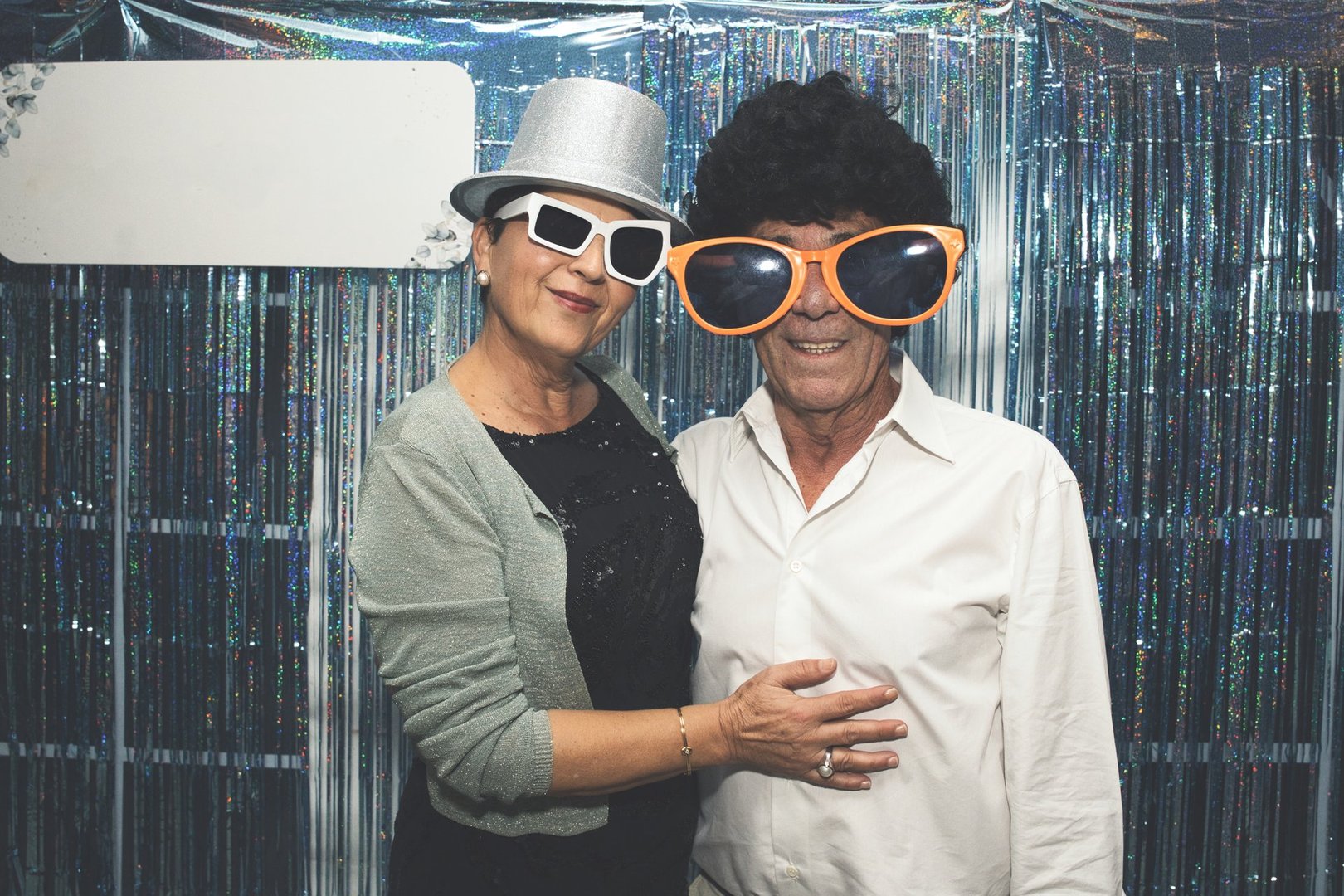 Senior couple wearing oversized glasses, a playful wig, and a fun hat, posing together for a photo booth, smiling joyfully and enjoying the lively atmosphere of a party