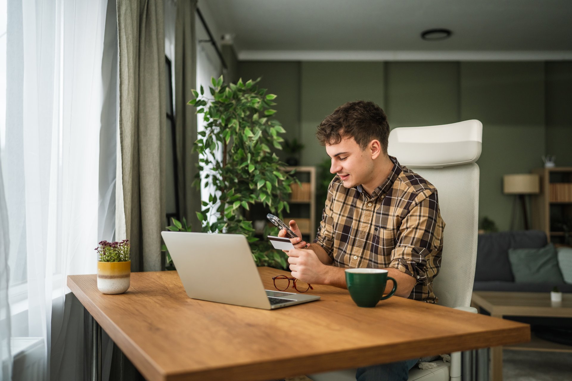 Young man sitting at a desk in a home office, making an online purchase using his smart phone and a credit card while a laptop, eyeglasses, and coffee mug are on the wooden surface