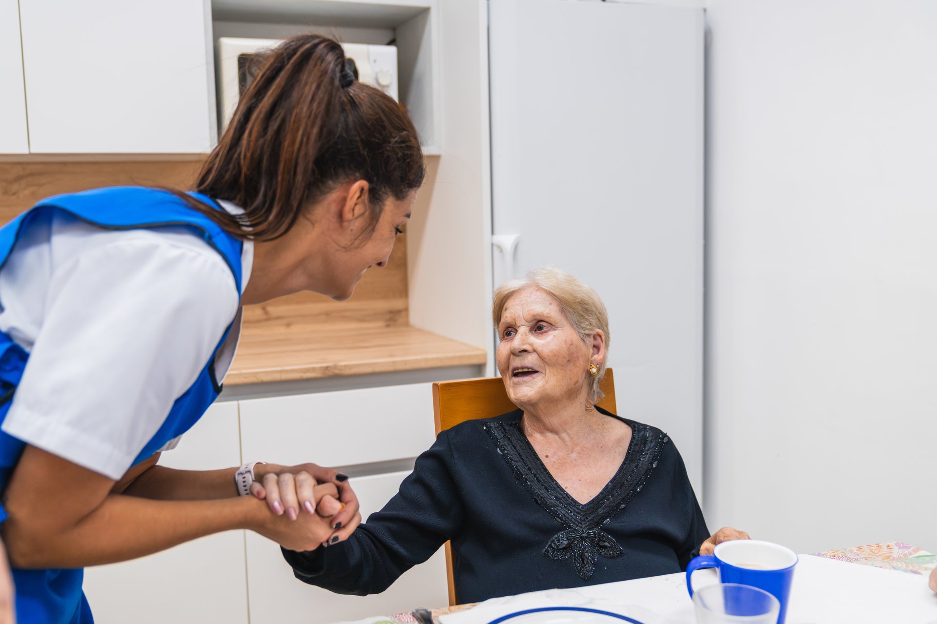 Nurse in blue uniform warmly engages with an elderly woman seated at a table, symbolizing compassionate care in a geriatric nursing home setting
