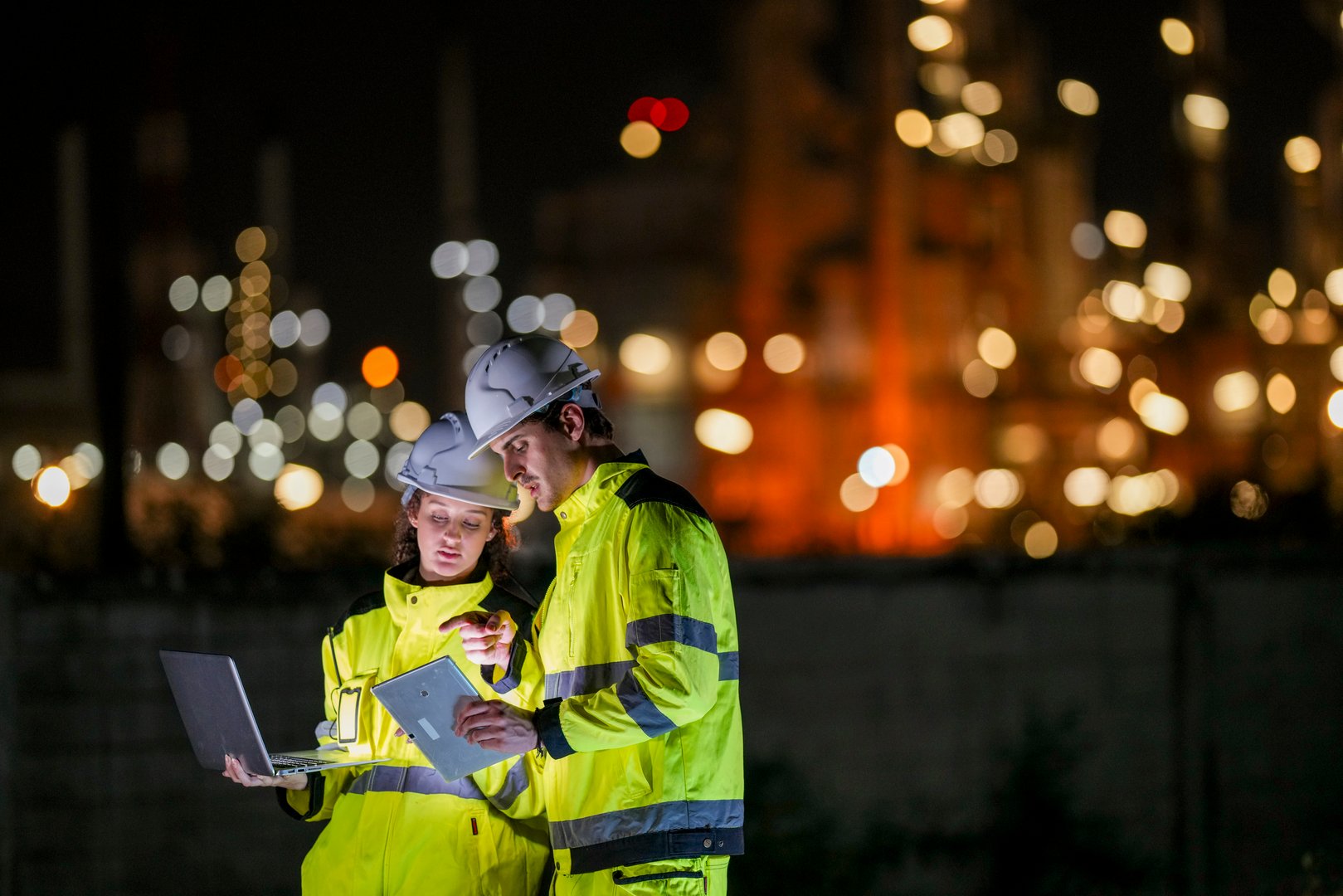 Two engineers wearing high-visibility suits review data on a laptop and tablet at night in front of a brightly lit industrial refinery, symbolizing night shift operations and teamwork.
