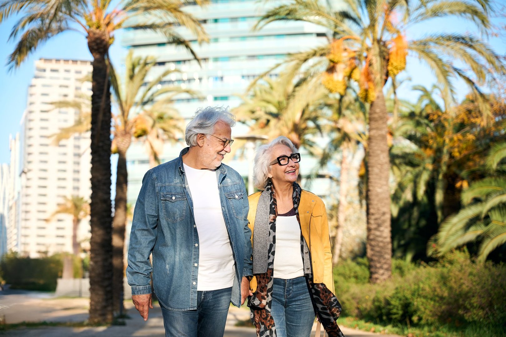 Cheerful retired couple enjoying a leisurely stroll through a vibrant city park adorned with palm trees, savoring the pleasant weather and each other's company during their golden years