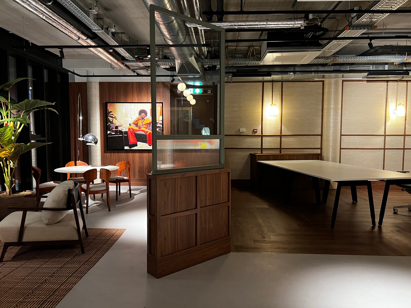 Modern office space with stylish furniture, a painting on the wall, and exposed ceiling pipes, softly lit by pendant lights.
