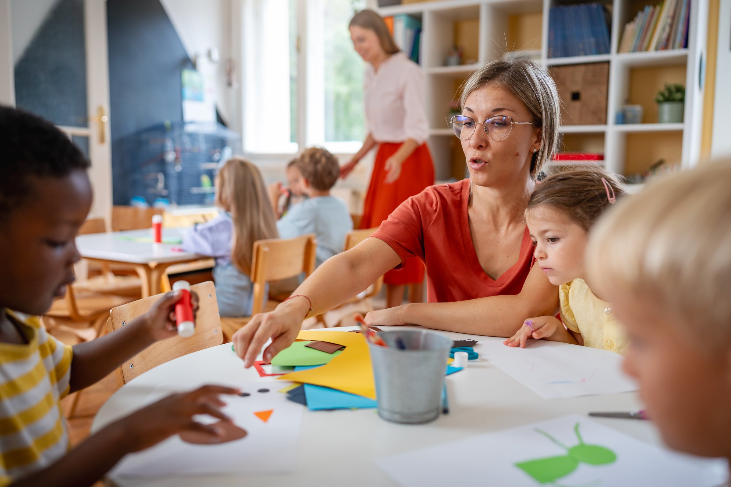 Preschool teacher assisting diverse children with arts and crafts in a bright, cheerful classroom