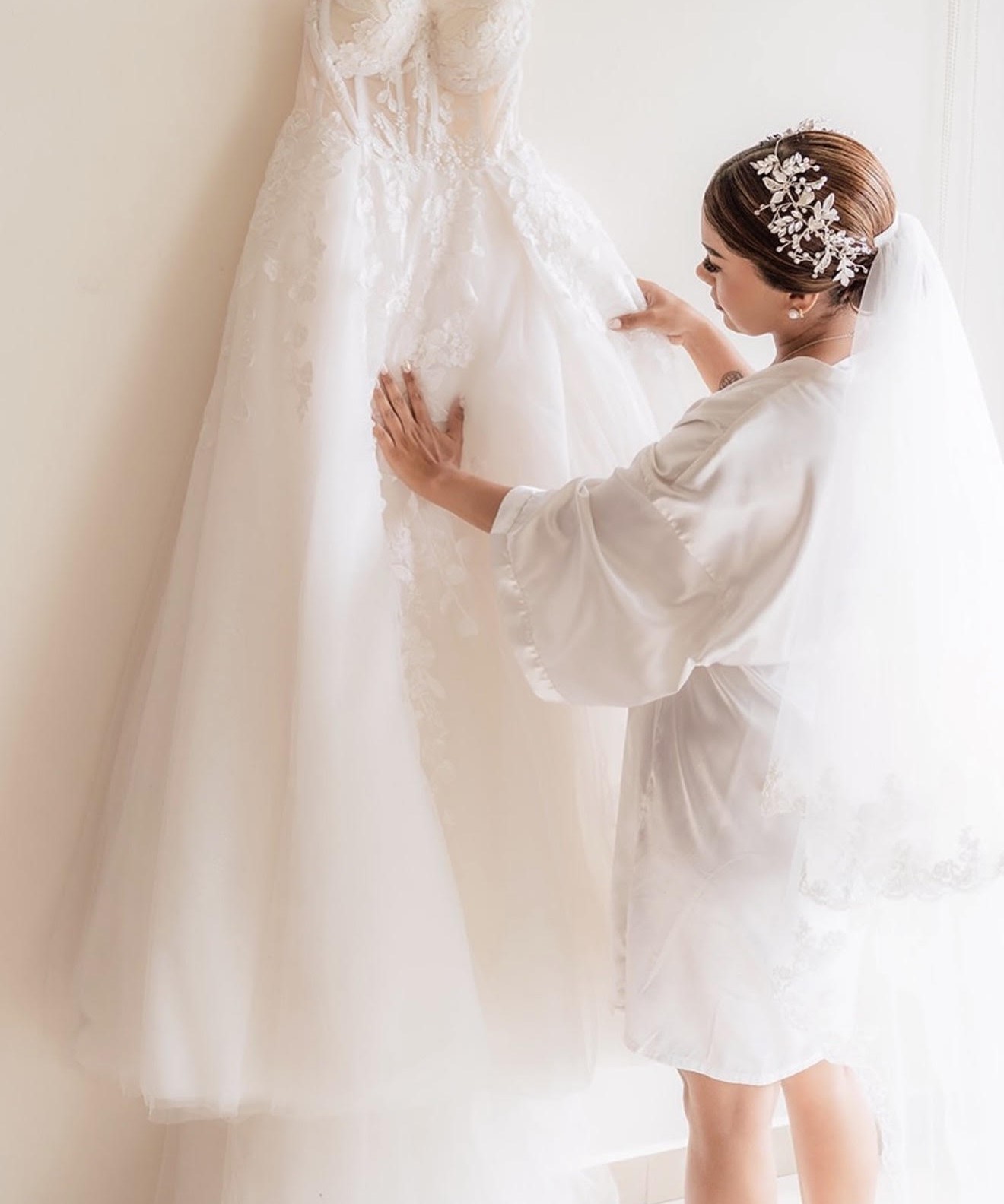 Bride in a robe admires her wedding dress hanging on a wall, preparing for her wedding day.