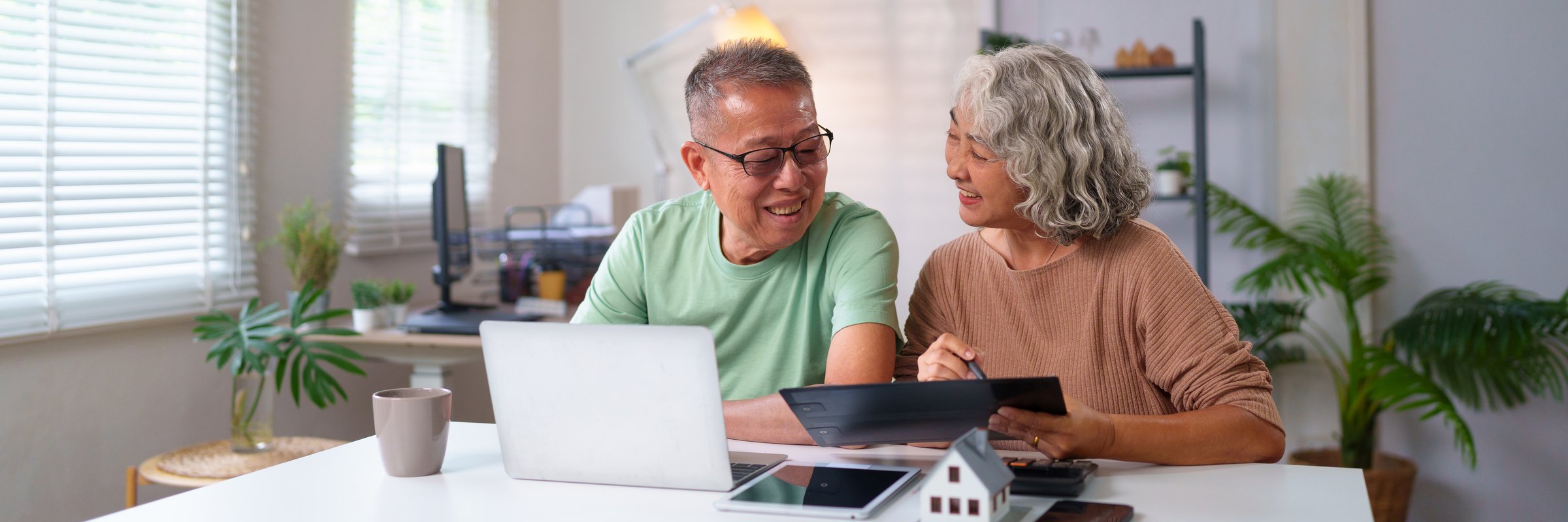 Happy older Asian couple smiling at home using tablet and laptop for retirement planning. Enjoying their life together in their cozy home with love.