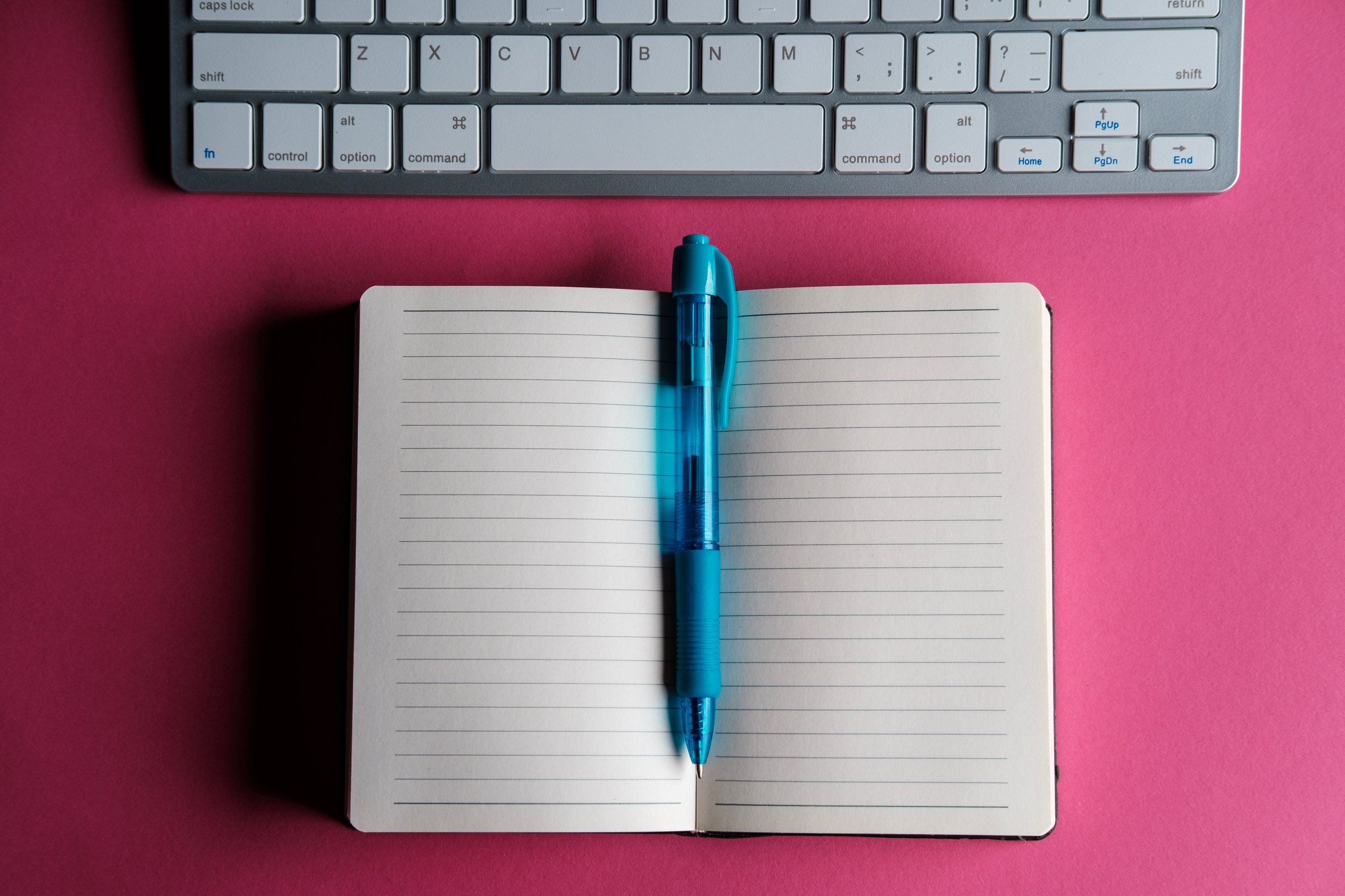 Open notebook with blank pages and blue pen lying on pink surface near wireless keyboard suggesting creative writing, note taking, or office work