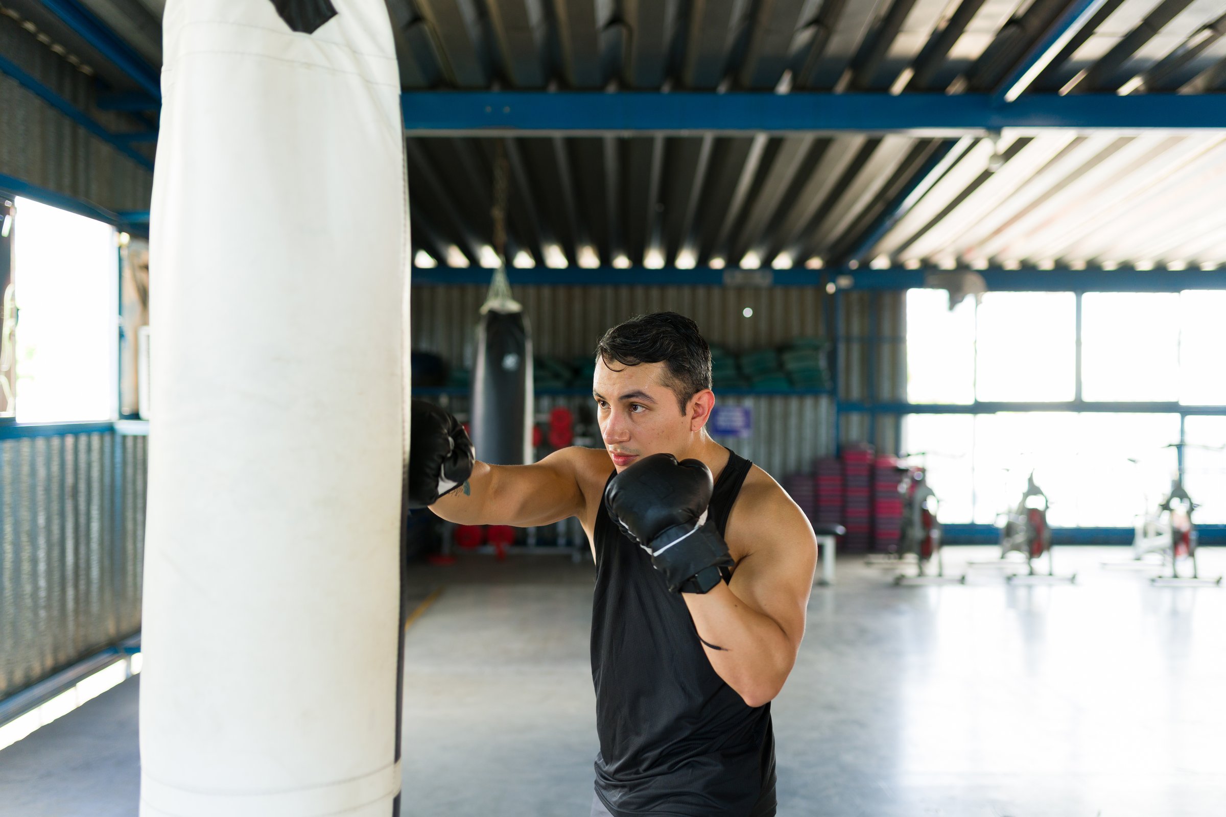 Young man wearing boxing gloves punching heavy bag during workout session. Focused on fitness, strength, and endurance