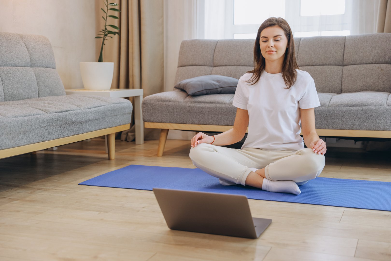 Woman sitting in lotus position on yoga mat, engaging in an online class with laptop, surrounded by a serene living room atmosphere