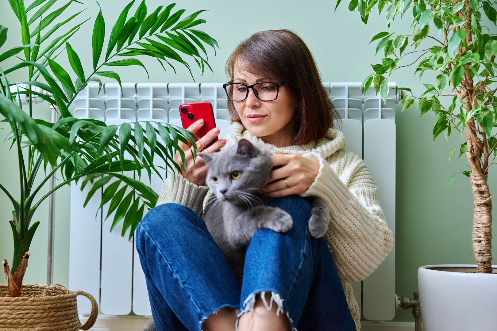 Middle aged woman resting at home in warm clothes using smartphone, warming herself with pet cat near home heating radiator. Winter autumn heating season, lifestyle leisure save energy, crisis concept