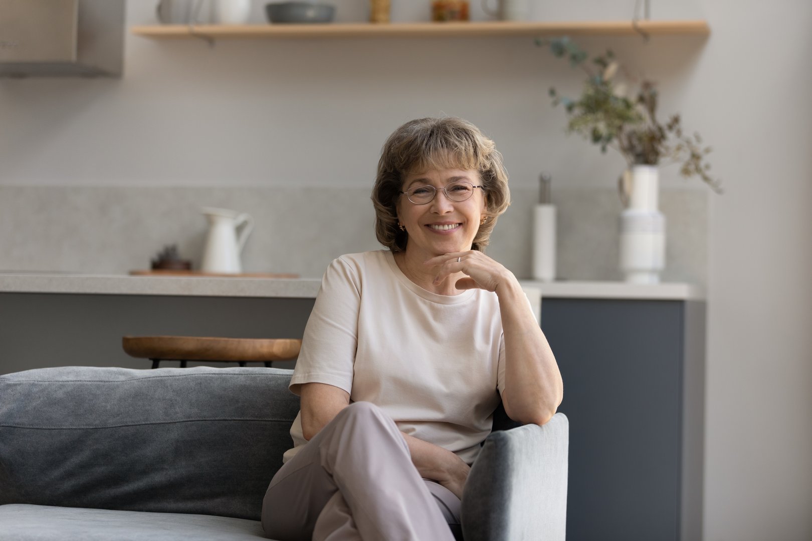 Portrait of smiling senior old retired woman in eyewear relaxing on comfortable sofa in living room. Happy peaceful middle aged mature grandmother enjoying peaceful weekend time, looking at camera.
