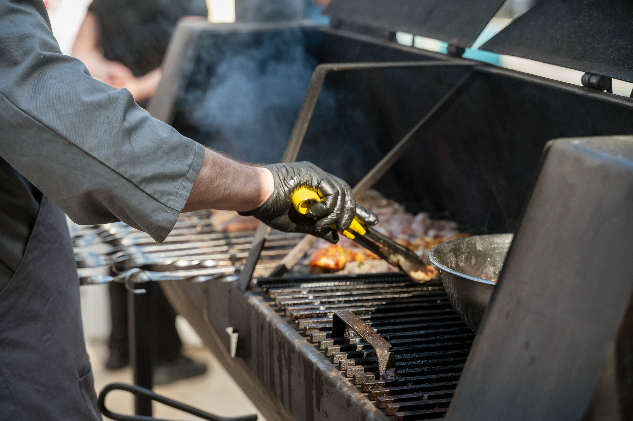 Male chef preparing meat on grill at barbecue party in summer garden, chef's hands in gloves. Concept of food, people and family time. Pork on skewers is roasted on coals in large metal grill device.