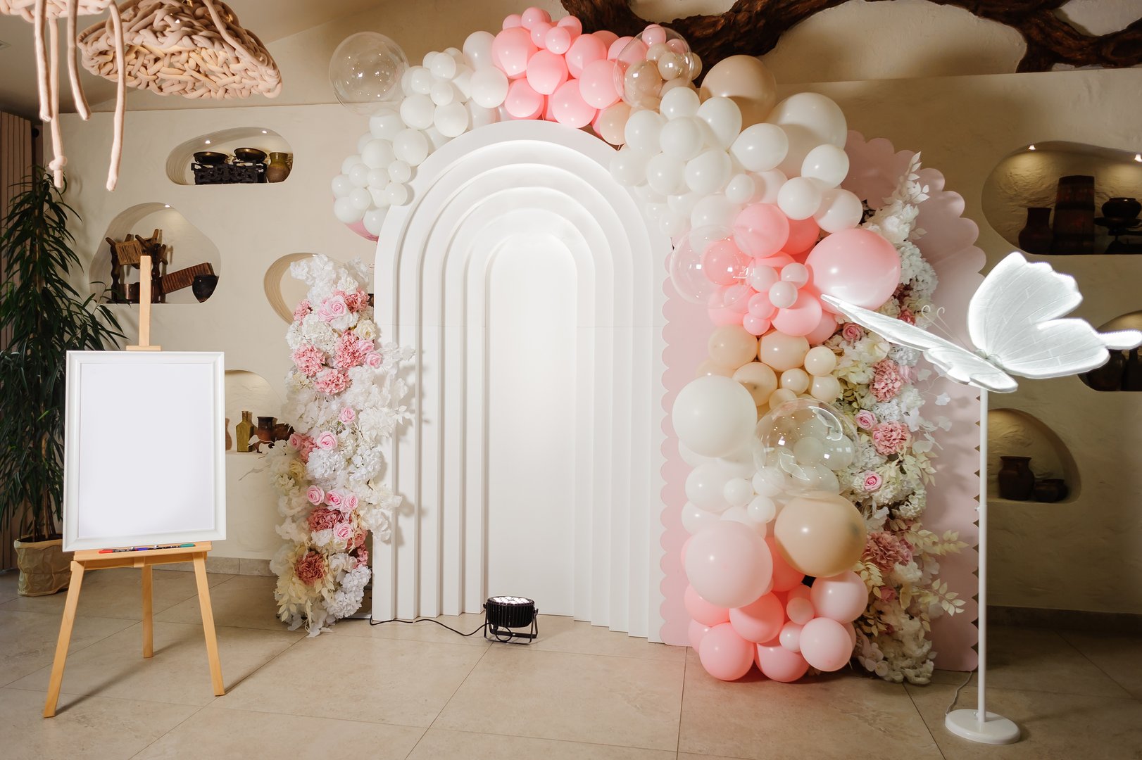 An elegant event photo zone featuring a modern white arch, a pink and white balloon garland, and a blank mockup easel, perfect for a wedding, baby shower, or birthday celebration