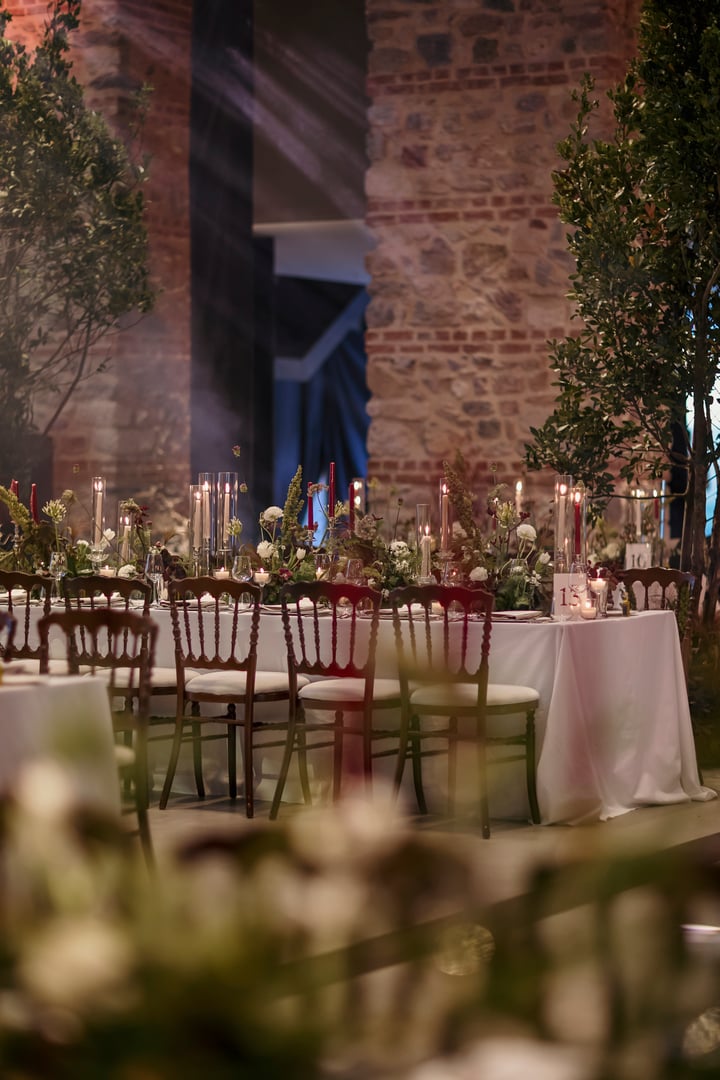 Beautifully decorated wedding reception hall featuring long banquet tables adorned with lush greenery, white flowers, and a mix of red and ivory candles. The setting is enhanced by classic wooden chairs, romantic candlelight, and a backdrop of rustic stone walls and arched windows. A sophisticated and atmospheric venue for a special celebration or event.