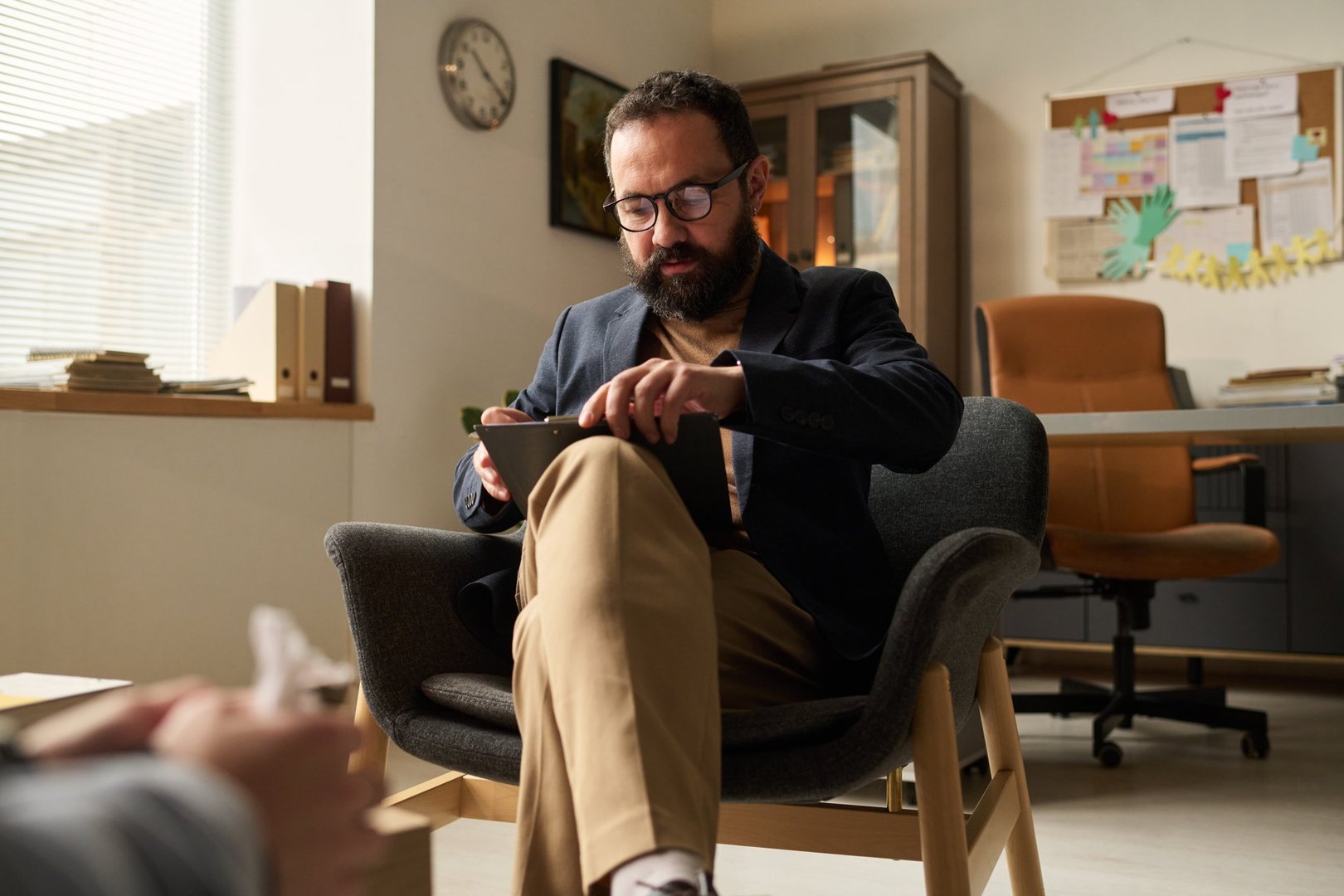 Serious school psychologist sitting in front of patient and looking through paper document on his knees in office