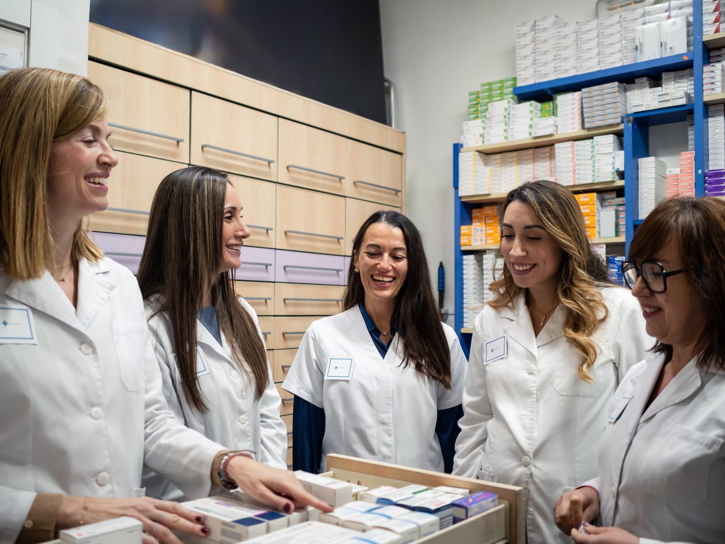 Inside a pharmacy, five women pharmacists are engaged in lively conversation while organizing medication. Their teamwork enhances the welcoming atmosphere as they work together.
