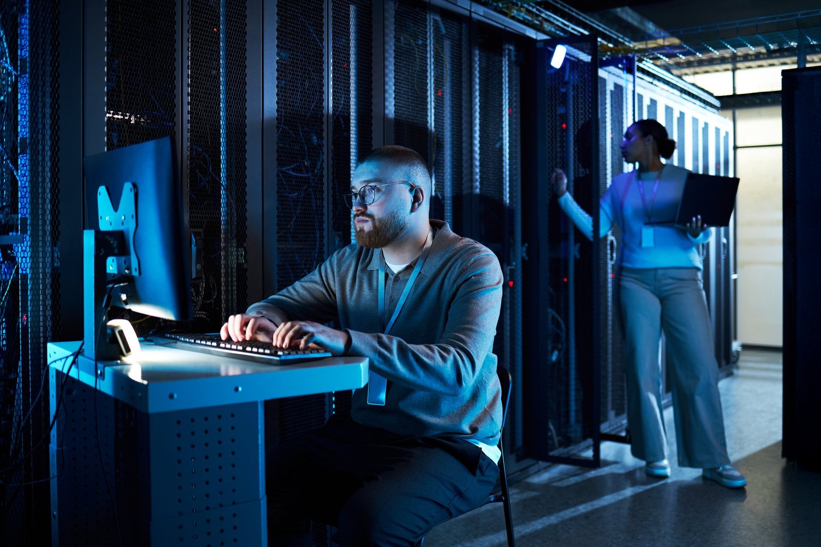 Technician monitoring server performance on computer while colleague analyzes data on tablet in secure server room environment with advanced technology and modern infrastructure