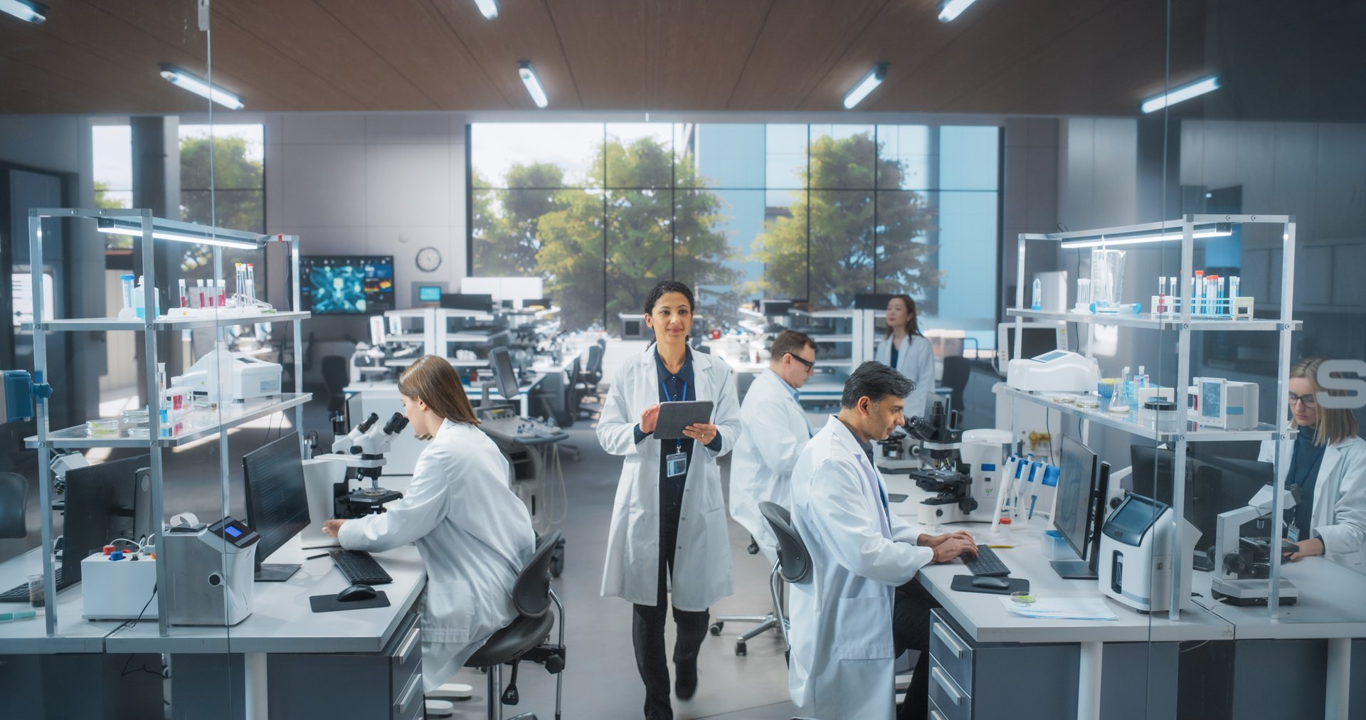 Team of Diverse Male and Female Medical Research Scientists Work on a New Generation Medical Products in a Modern Laboratory. Female Walking with Tablet Computer, Browsing Research Data