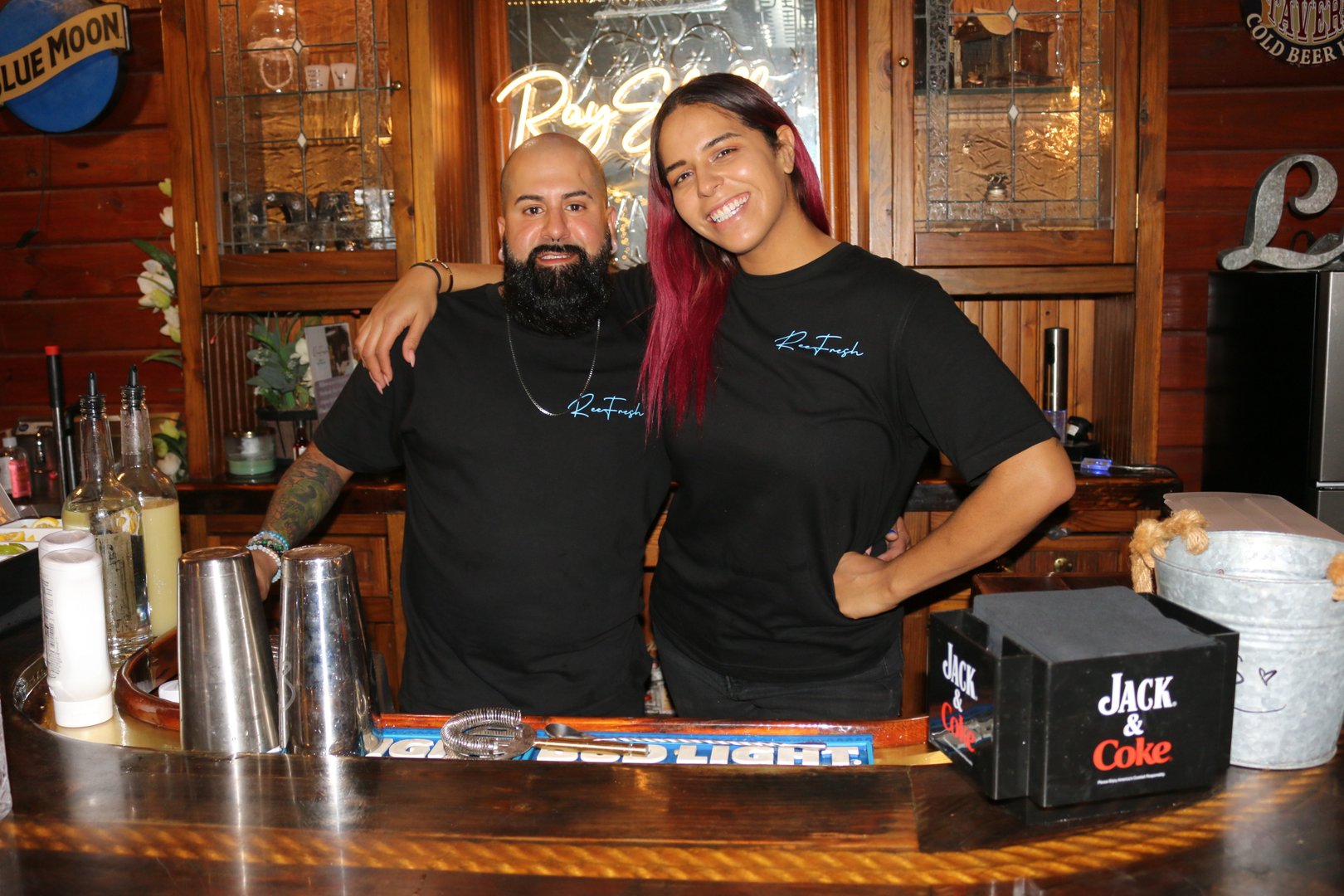 Two people smiling behind a bar, wearing matching black shirts. Liquor bottles and bar tools are visible on the counter.